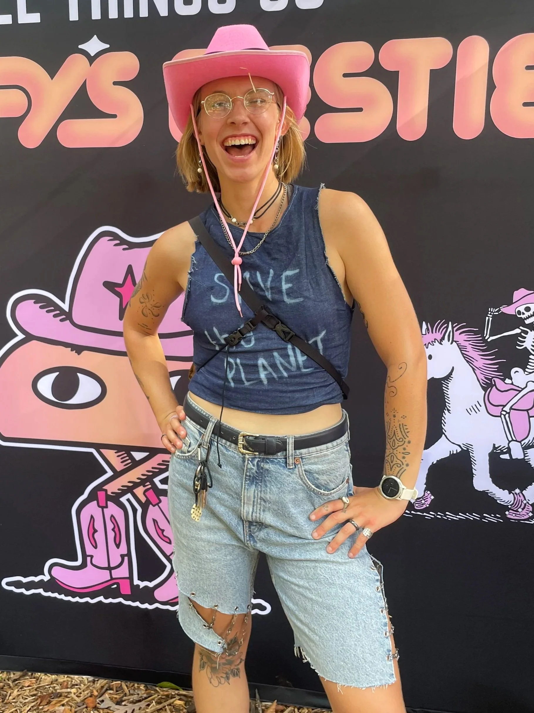 A woman wearing a pink cowboy hat, glasses, and a sleeveless denim top with the words 'Save the Planet' stands in front of a black backdrop with cartoonish pink and white illustrations of a cowboy hat, a cat, a unicorn, and a skeleton riding a horse. She is smiling broadly and has tattoos on her right arm and left leg.