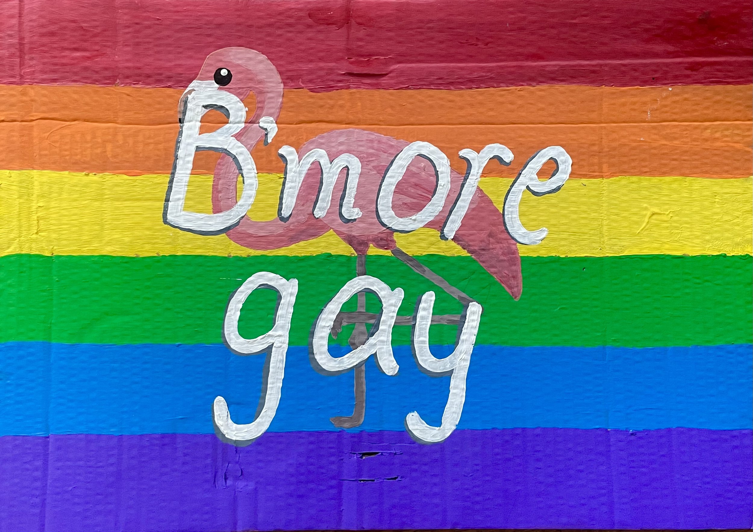 A colorful mural featuring horizontal rainbow stripes with a pink flamingo in the center. Overlaid on the image are the words 'B'more Gay' in white with a black outline, with the flamingo's body and leg integrated into the text.