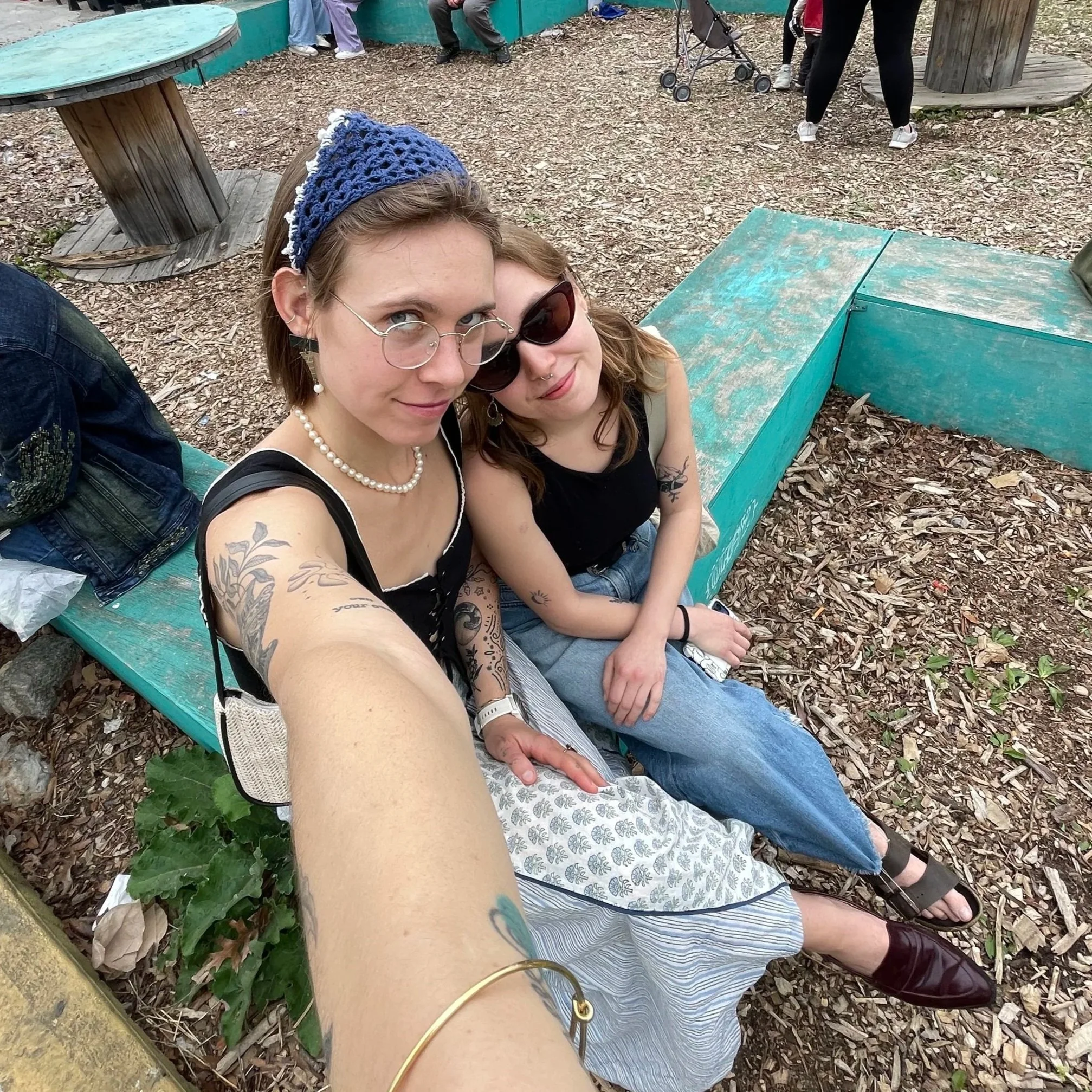 Two young women sitting on a green bench outdoors, taking a selfie. They are wearing sunglasses, one with a pearl necklace, and the other with a nose ring. They are surrounded by wood chips and greenery, and there are other people in the background.