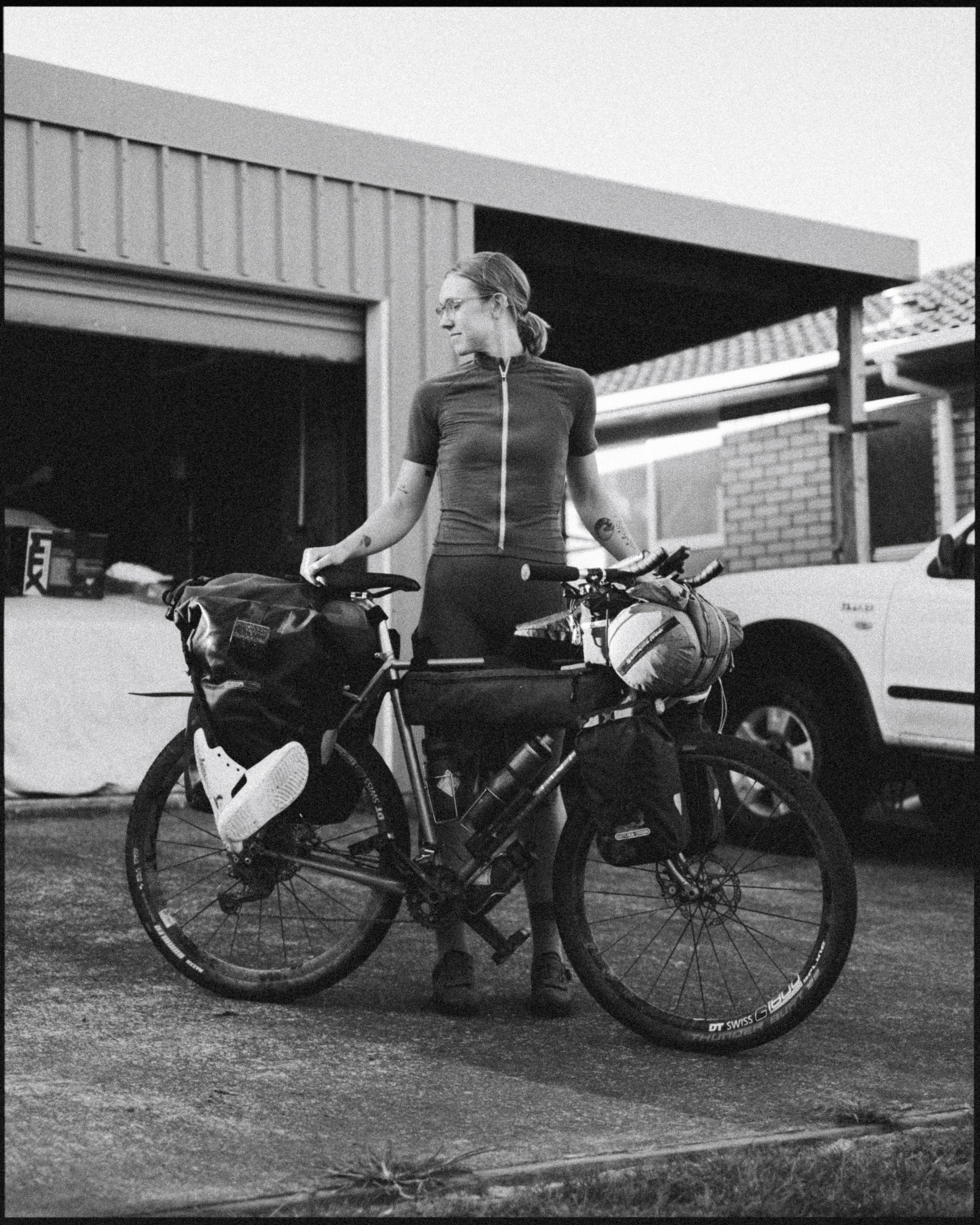 A woman with glasses standing with a loaded touring bicycle in front of a garage.