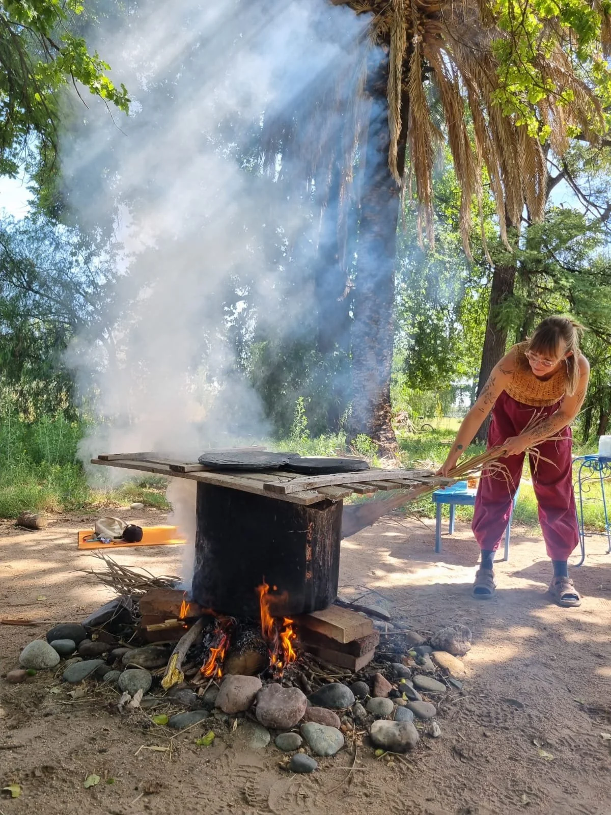 A person with tattoos, wearing glasses, a sleeveless top, and maroon pants is tending to a fire under a makeshift outdoor cooking setup. The fire is burning on the ground surrounded by rocks and is heating a large black pot placed on a wooden platform. Smoke is rising from the fire, and the scene is set outdoors under trees with green foliage.