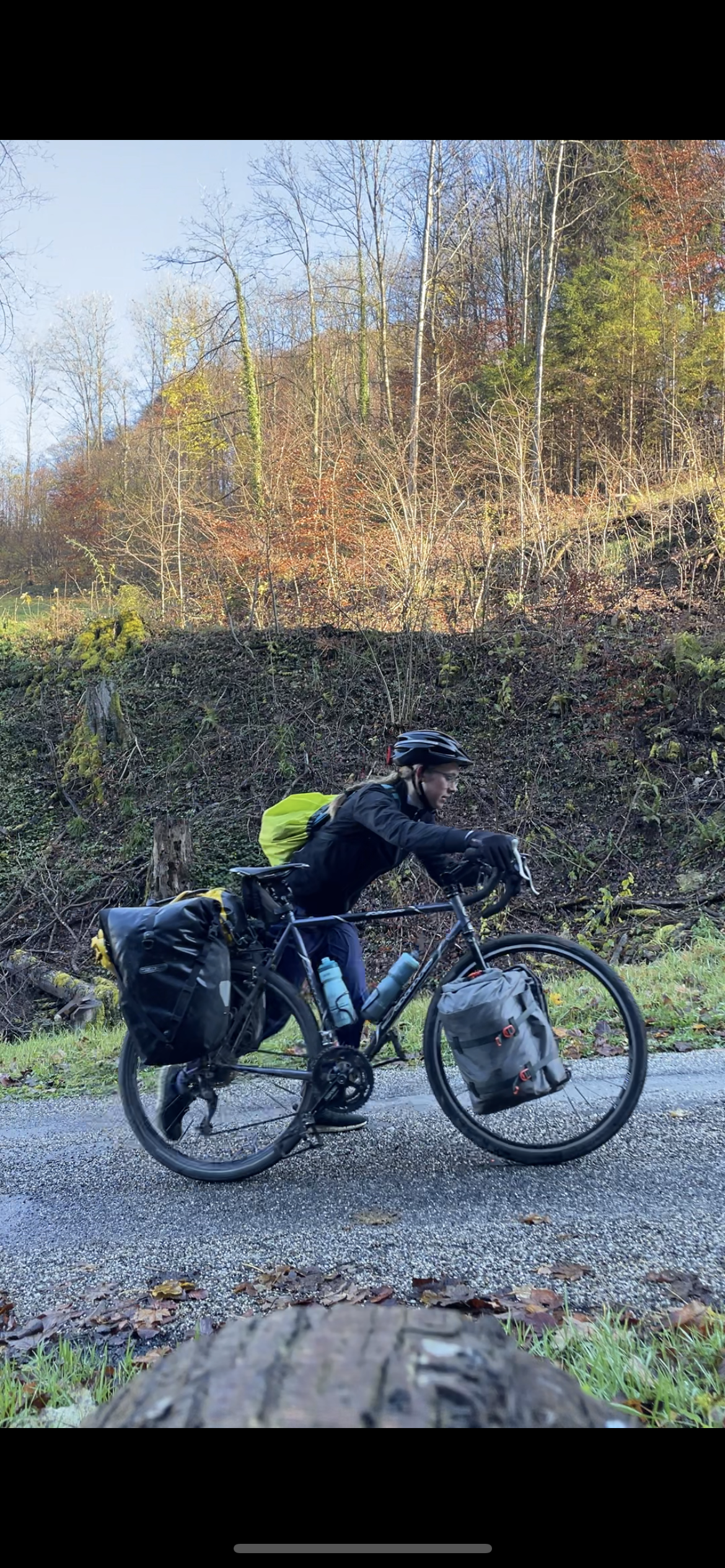A woman riding a loaded bicycle on a rural road with autumn trees in the background.