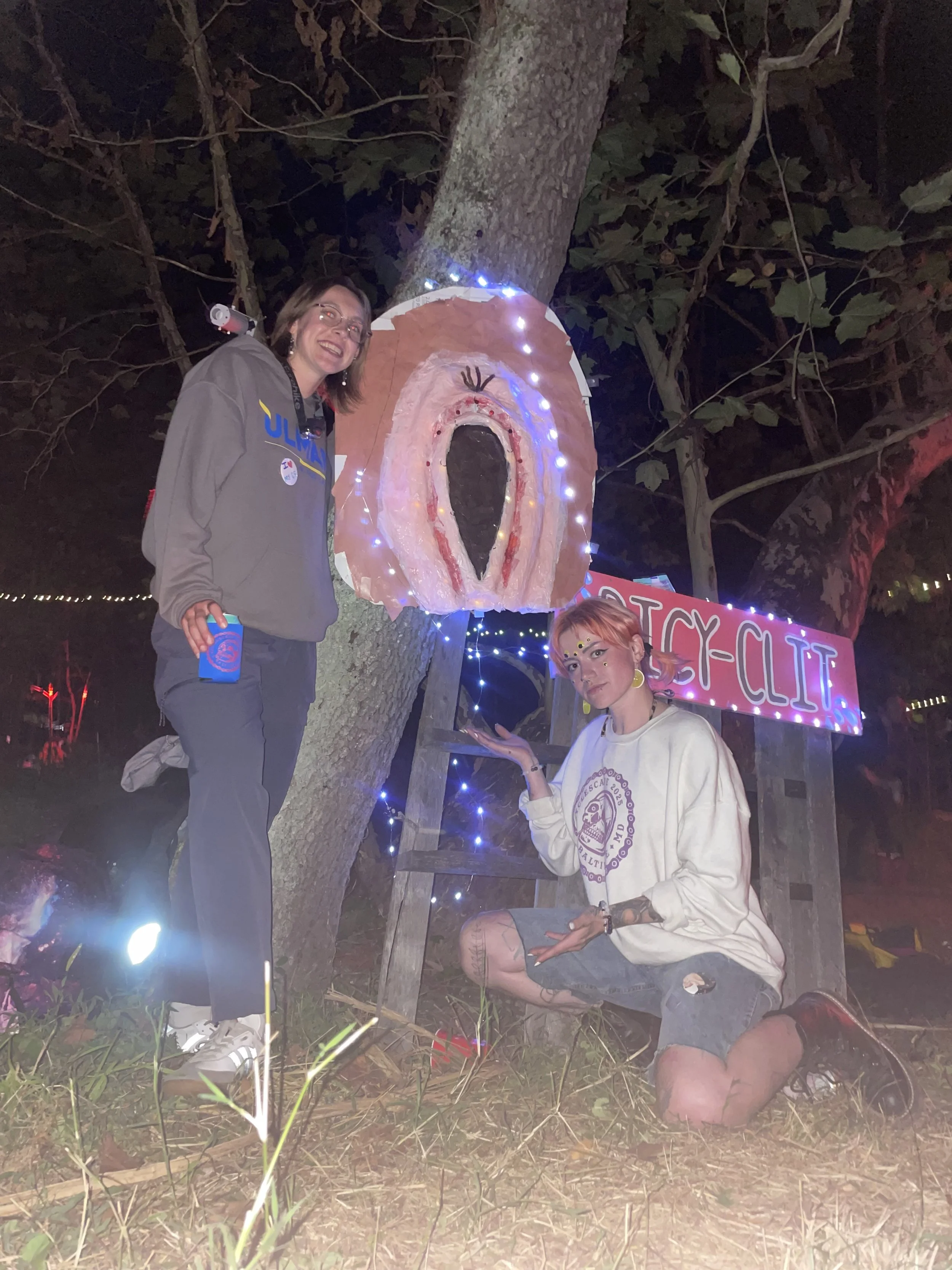Two young women at a Halloween-themed event at night, standing and kneeling in front of a tree with a painted face and tongue decoration, lit with string lights. One woman is smiling and holding a cup. The other woman has colorful makeup, tattoos, and is squatting, pointing to the decoration, with a sign behind that reads "TICKY-CIT".