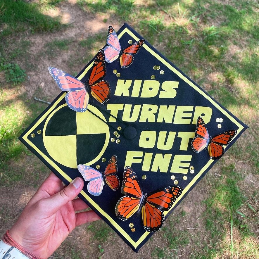 Graduation cap decorated with butterflies, gold sequins, and the phrase 'Kids Turned Out Fine' in yellow letters, held outdoors on grass.