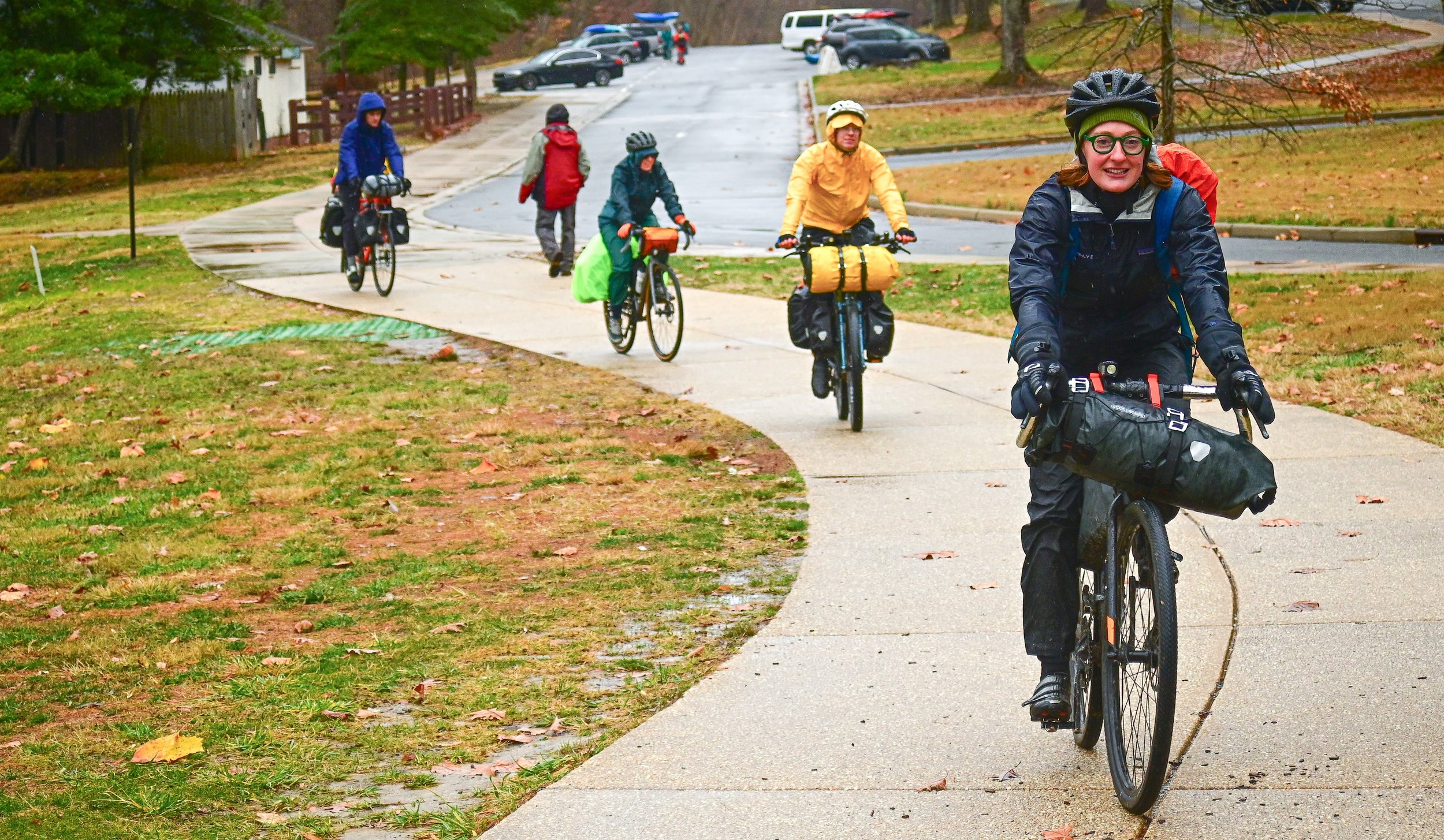 A group of five cyclists riding bicycles on a paved sidewalk in a neighborhood during a rainy day. They are dressed in rain gear and carrying gear bags, with trees and parked cars in the background.