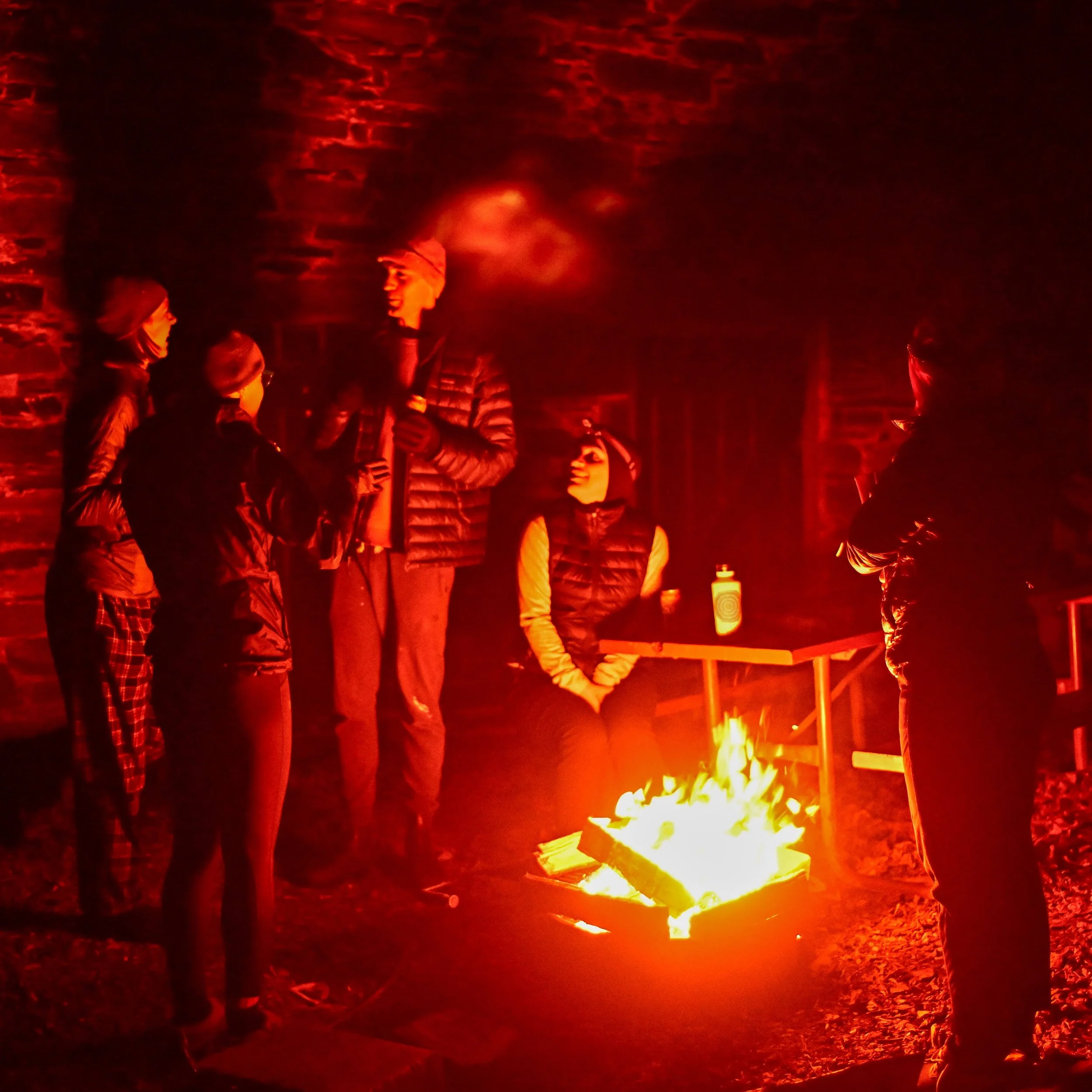 Group of people gathered around a campfire at night, some standing and some sitting, in a dark outdoor setting with brick wall in background.