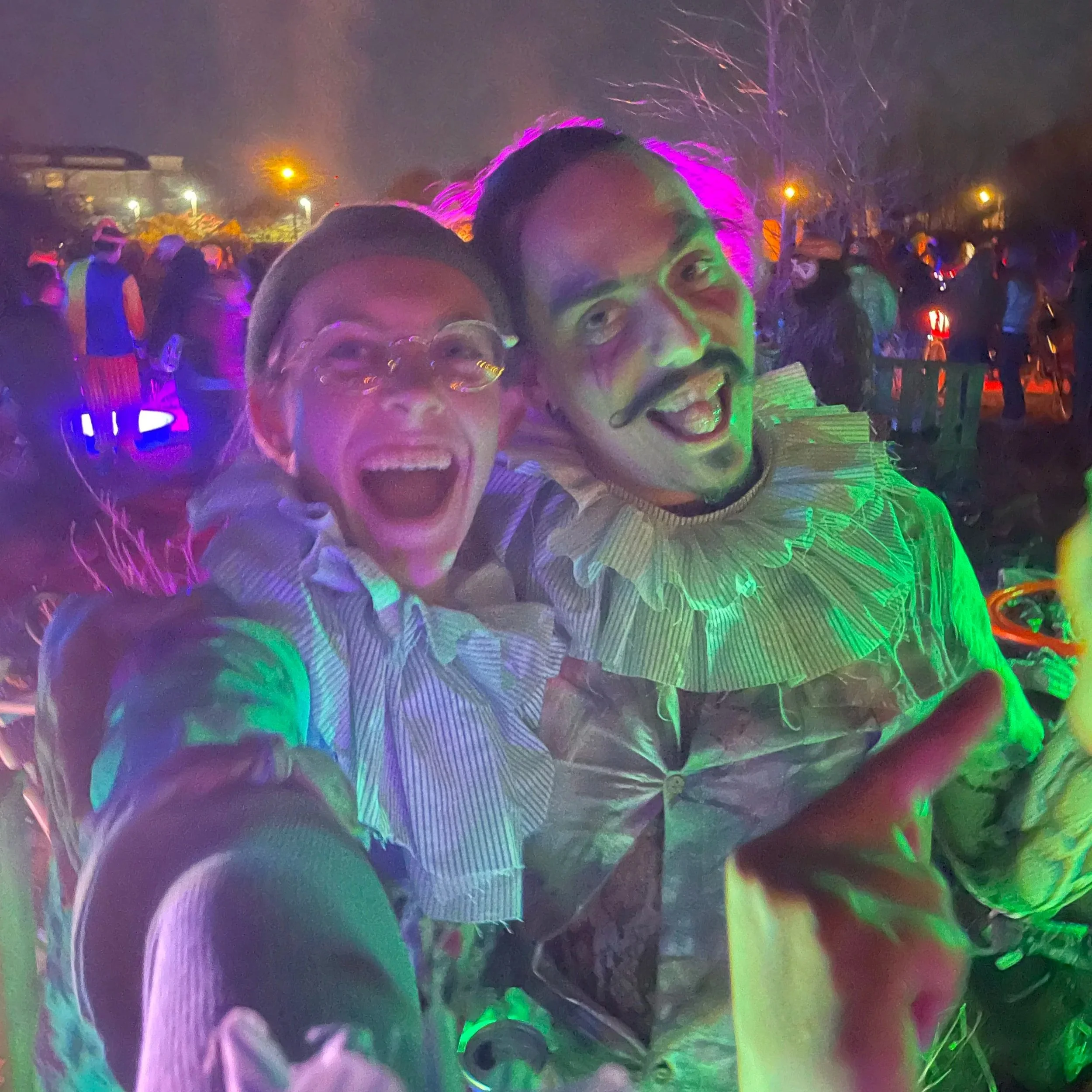 Two people dressed in colorful costumes, smiling and taking a selfie at a lively outdoor event during nighttime, with other people and bright lights in the background.