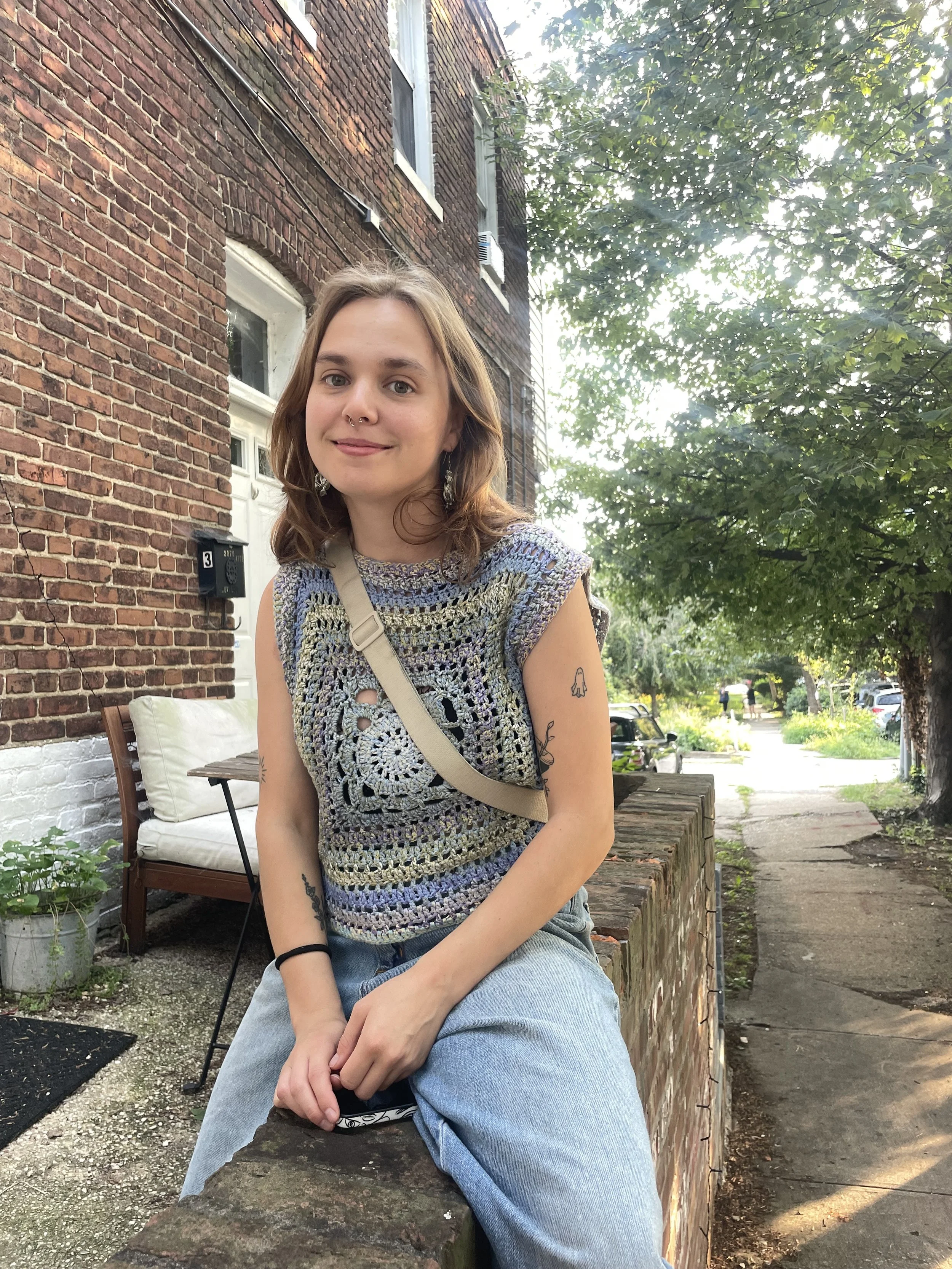 A young woman with shoulder-length brown hair and light skin sitting on a brick ledge outdoors, wearing a multicolored crocheted top and light blue jeans, with tattoos on her arms and a beige shoulder bag, smiling at the camera.