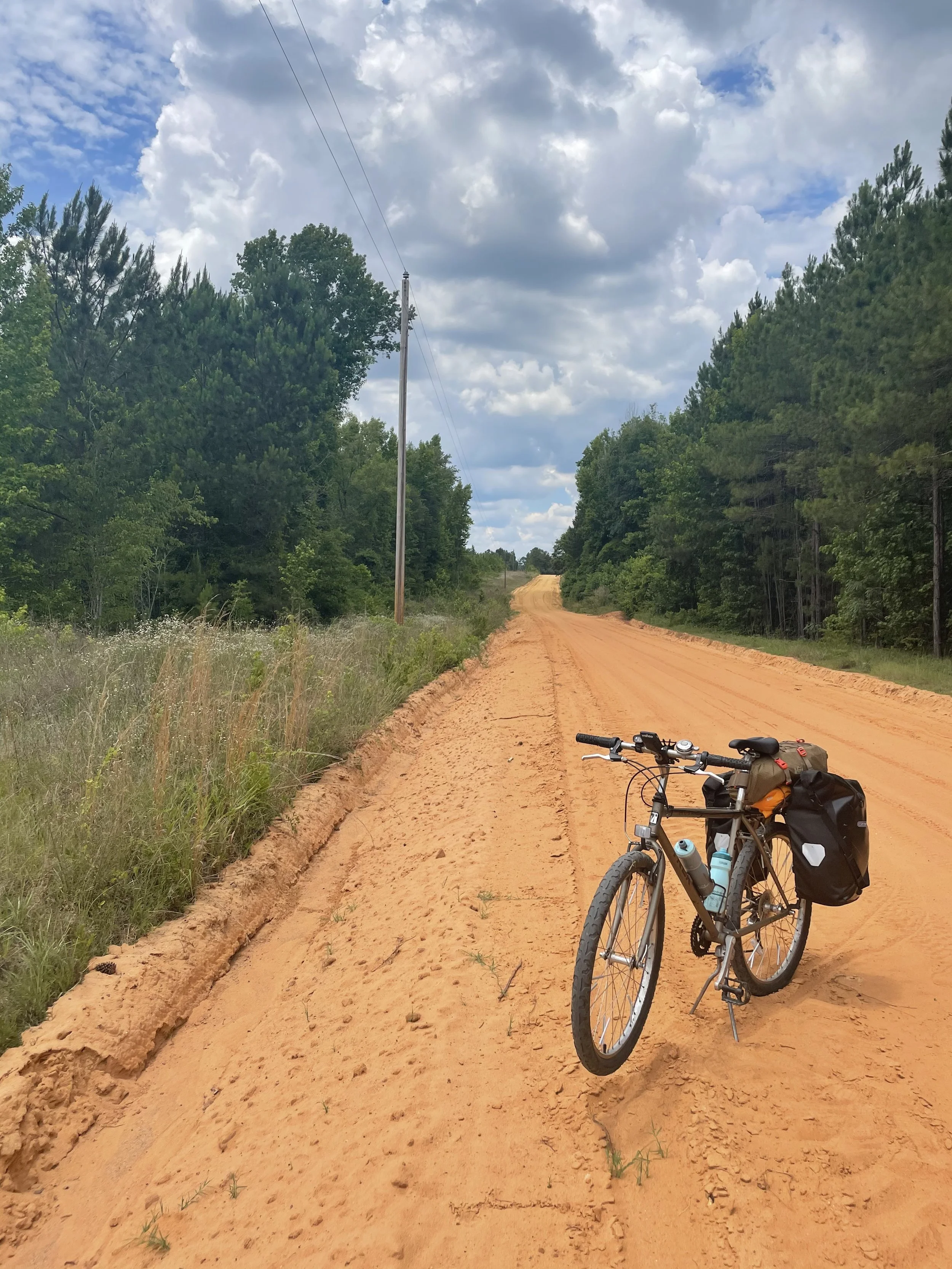 A mountain bike with a water bottle, dry bag, and saddlebag parked on a dirt road surrounded by green trees under a partly cloudy sky.
