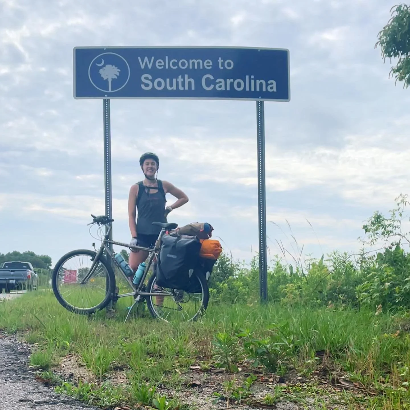 A smiling woman in cycling gear standing next to her bicycle loaded with gear, beneath a Sign that says 'Welcome to South Carolina'.