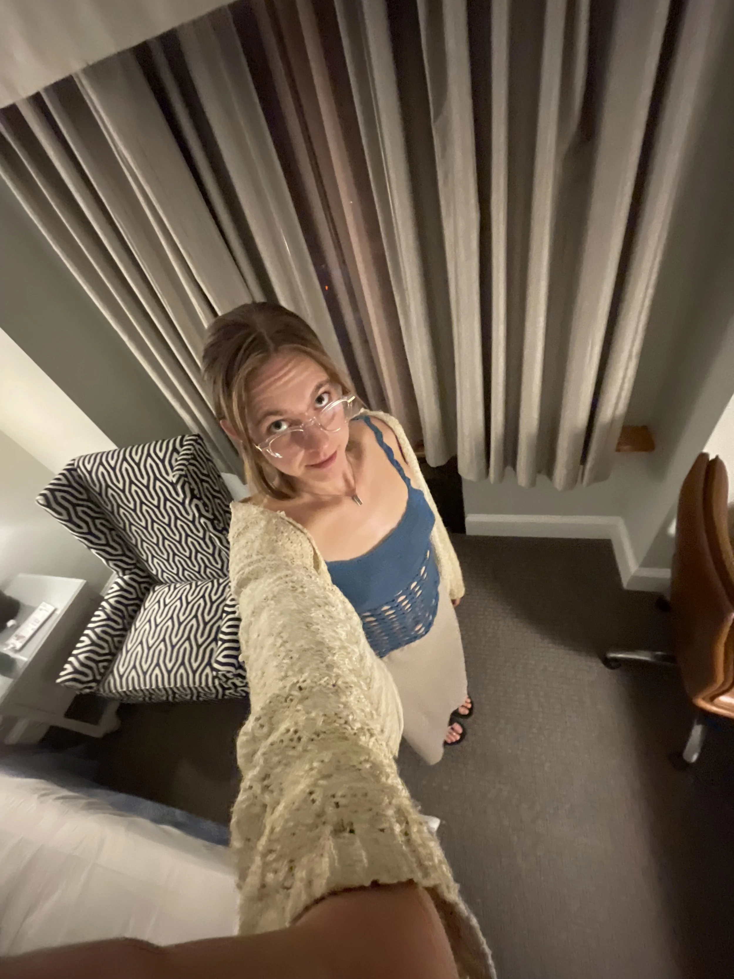 A woman with glasses and shoulder-length hair taking a selfie in a hotel room, looking up at the camera. She is wearing a blue top, a beige cardigan, and a beige skirt. Behind her are curtains, a bed with a patterned pillow, a nightstand, and a chair.