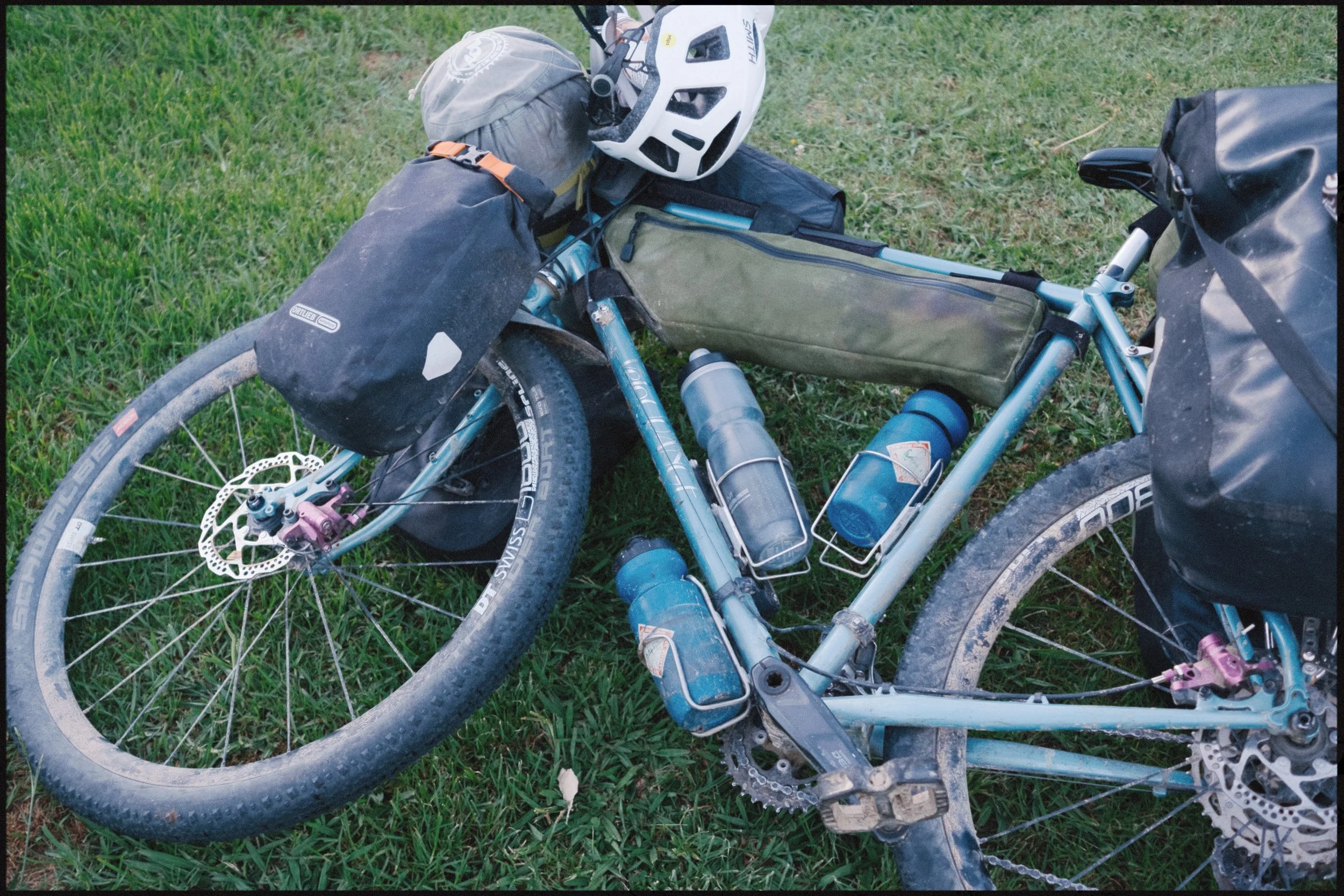 A blue mountain bike lying on grass, equipped with black panniers, a gray helmet, and multiple water bottles attached to the frame.