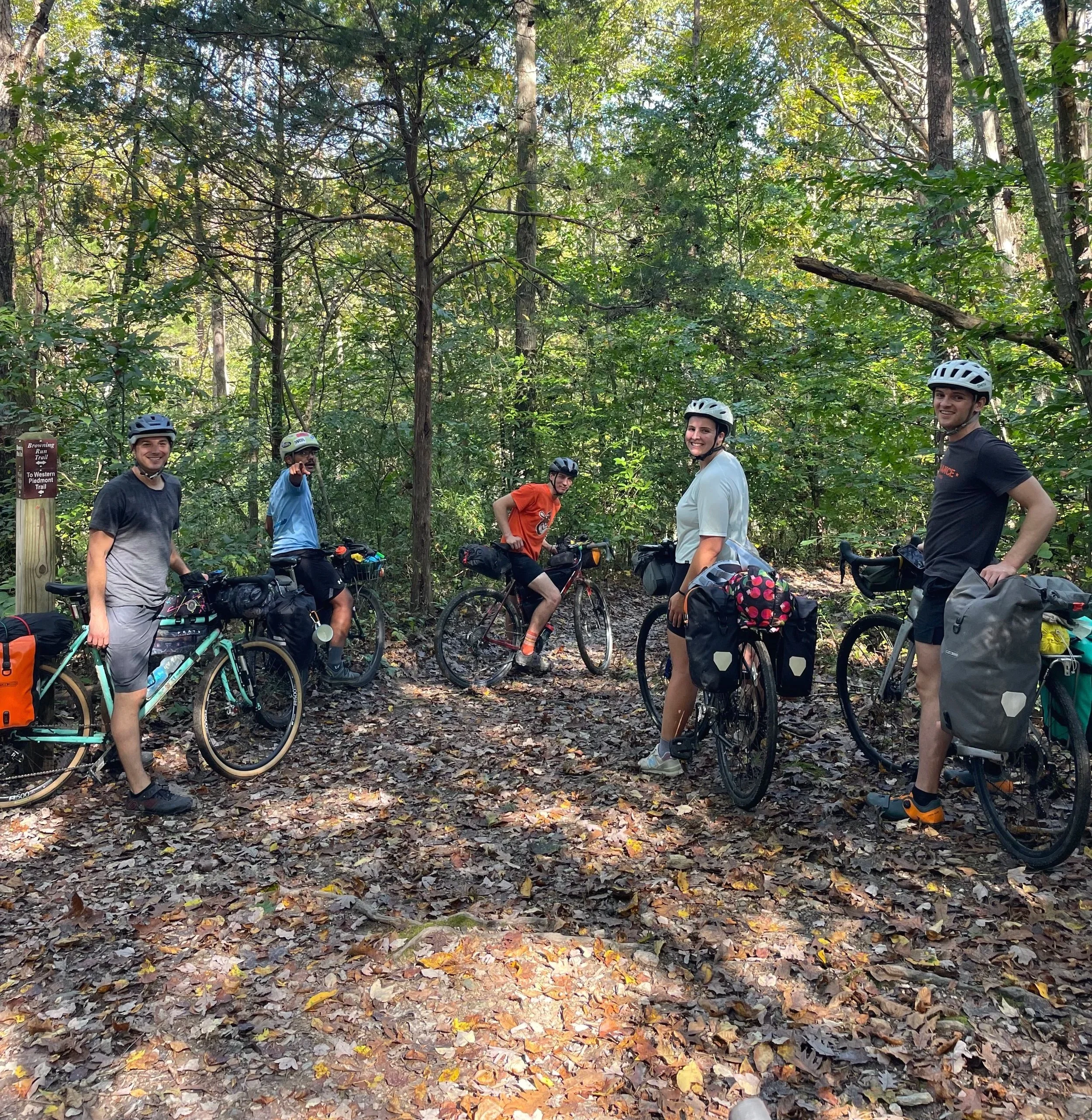 Five cyclists wearing helmets stand with their bikes in a wooded area, smiling and posing for the photo.