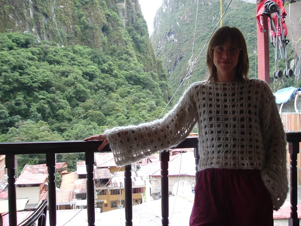 A woman standing on a balcony with a lush, green mountain landscape in the background. She is wearing a cream-colored, open-knit sweater and has shoulder-length hair, glasses, and a slight smile.