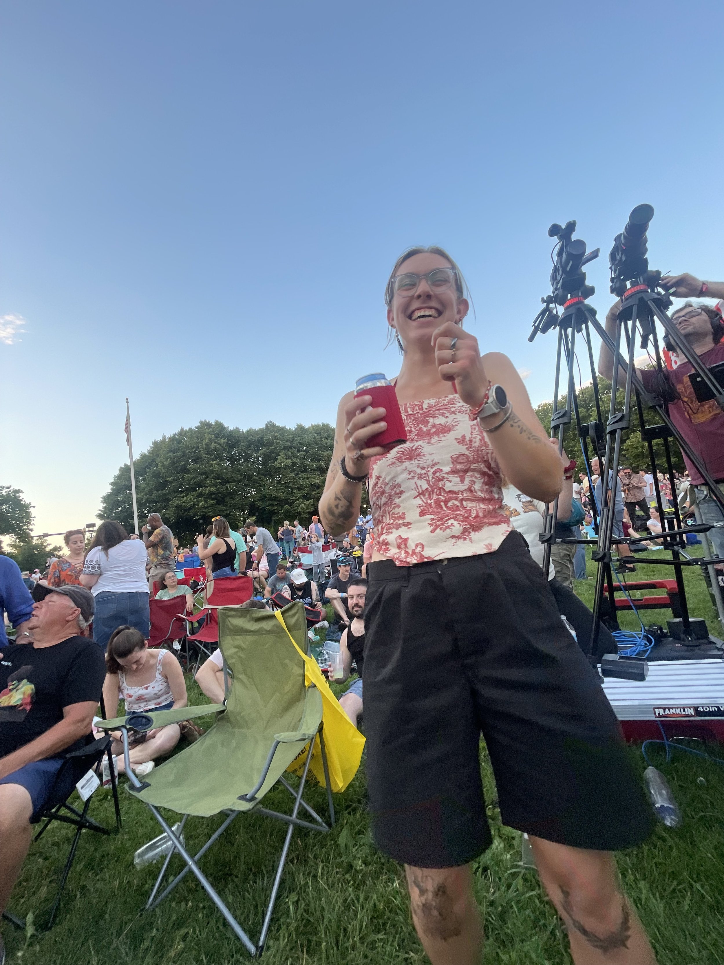 A woman with glasses and tattoos smiling and holding a drink at an outdoor event with a crowd, chairs, and camera equipment in the background.