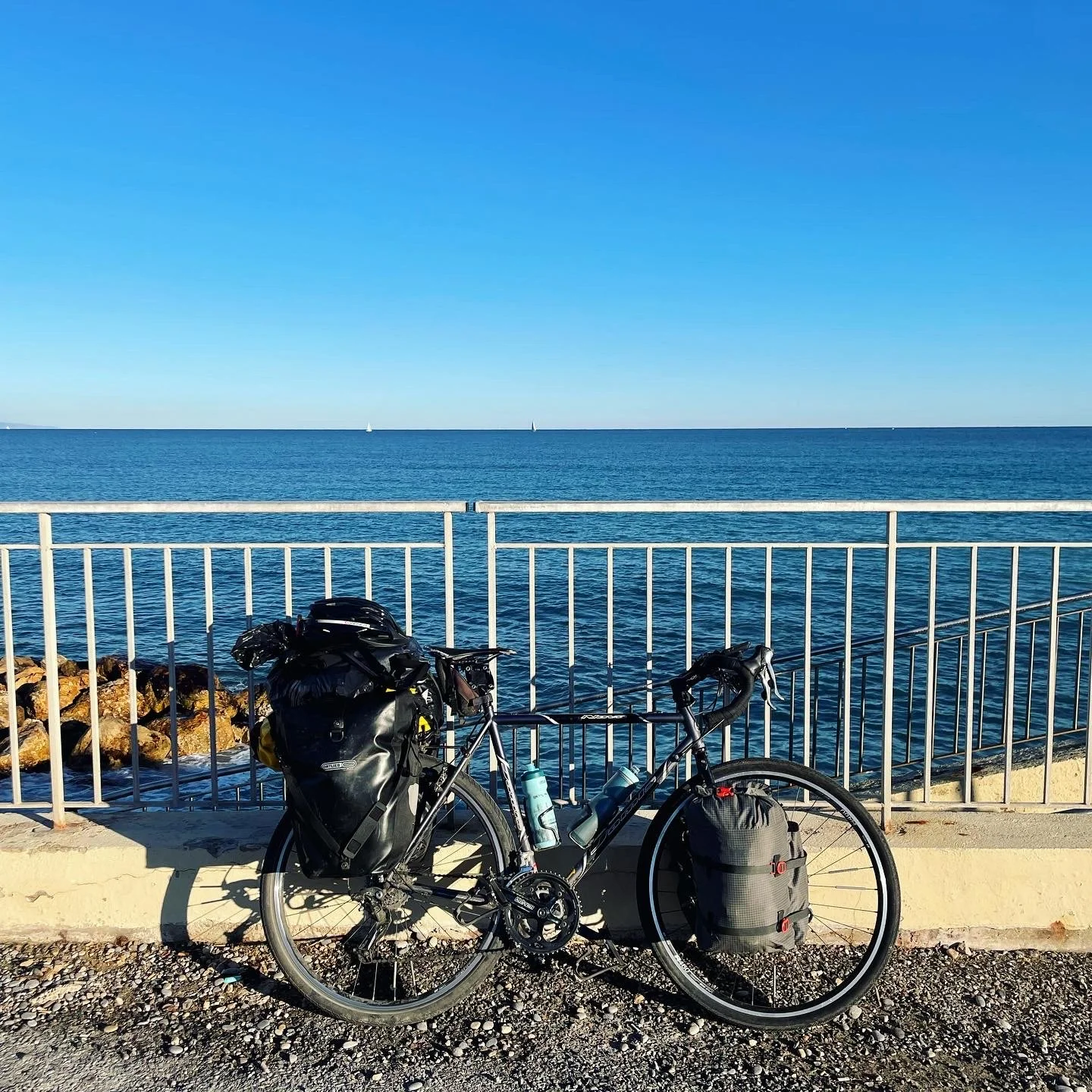 A bicycle with panniers and water bottles parked by a seaside railing, overlooking the ocean with a clear blue sky.