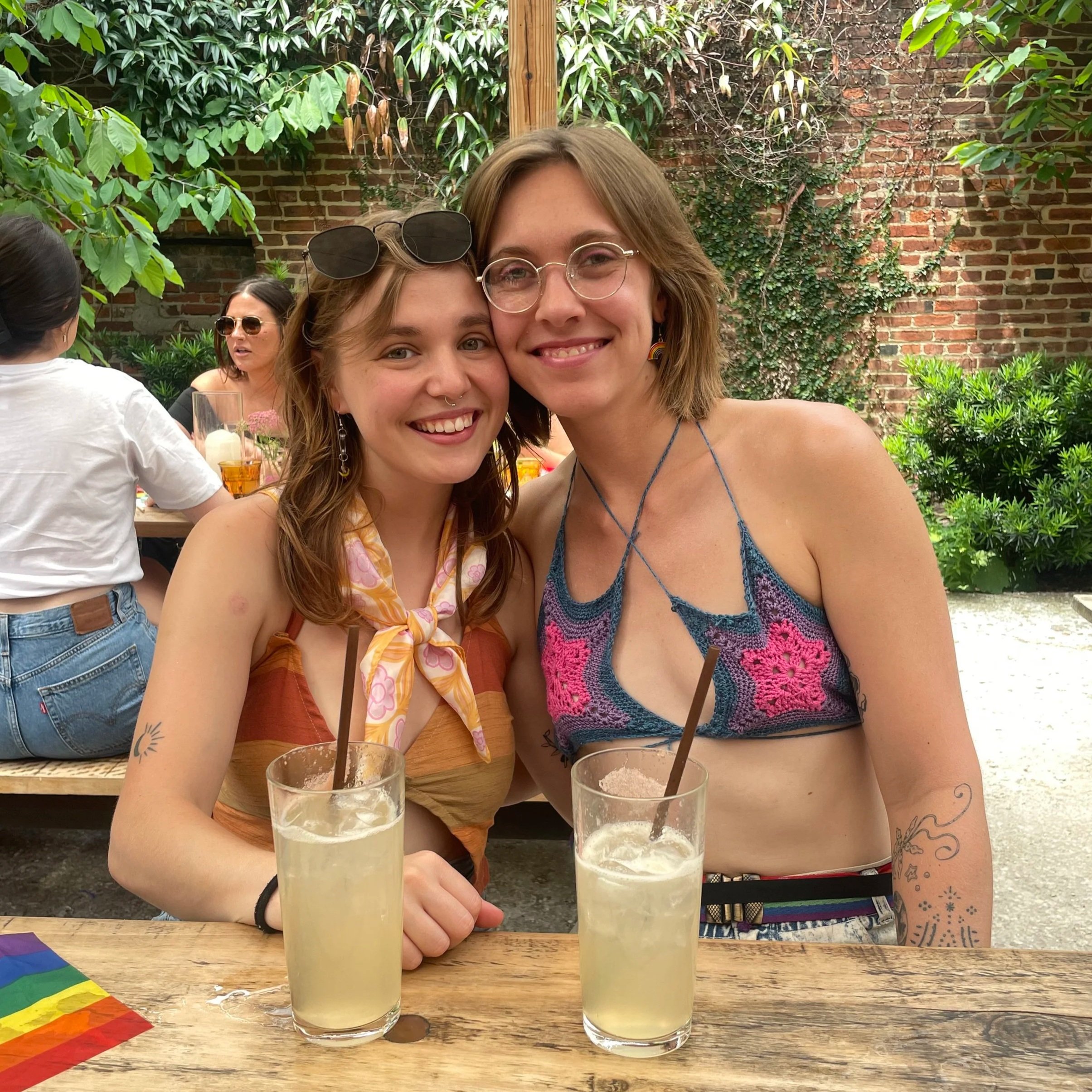 Two young women sitting at a wooden table outdoors, smiling and posing for the photo, with two drinks in front of them, surrounded by greenery and a brick wall in the background.