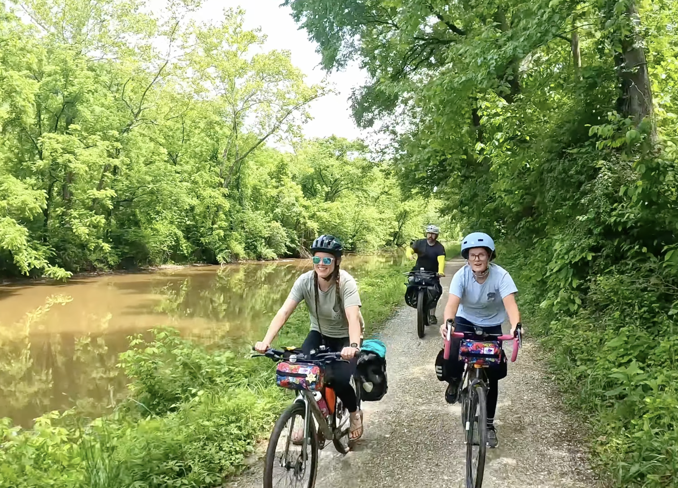 Three women and one man riding bicycles on a forested trail beside a river, all wearing helmets and smiling, with lush green trees surrounding them.