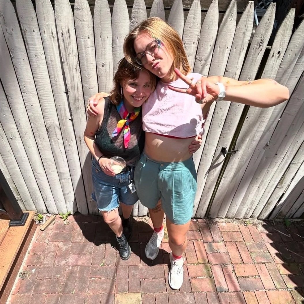 Two young women standing close together in front of a wooden fence on a brick patio, smiling and making peace signs. One is holding a drink and has a colorful scarf around her neck, while the other is wearing glasses and a pink cropped t-shirt.