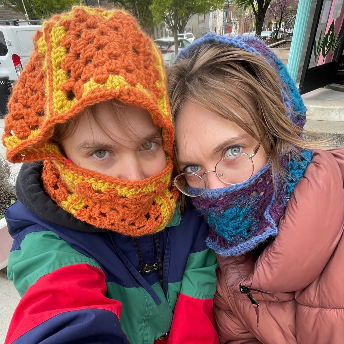 Two children bundled up in colorful, cozy winter hats and face coverings, taking a close-up selfie outdoors with trees and parked cars in the background.