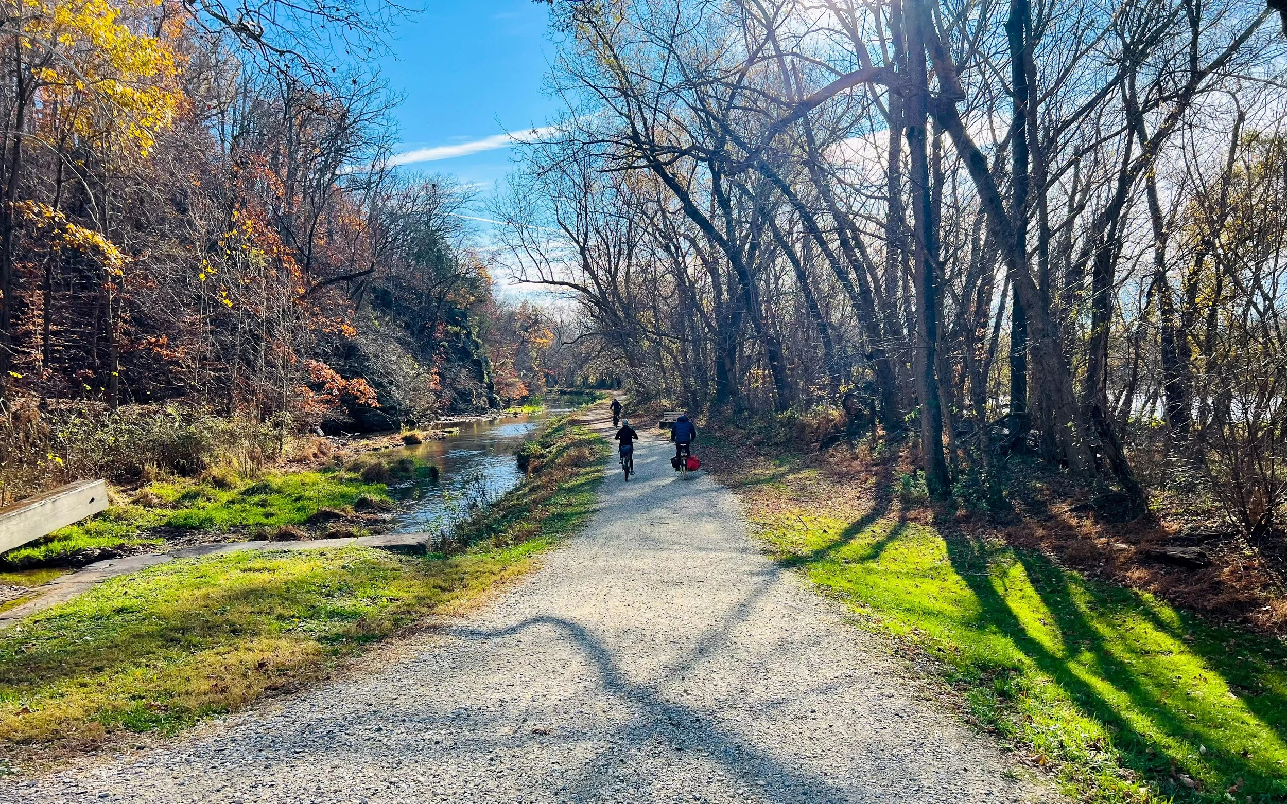 People riding bicycles along a gravel path by a small river or creek in a wooded park during fall or early winter.