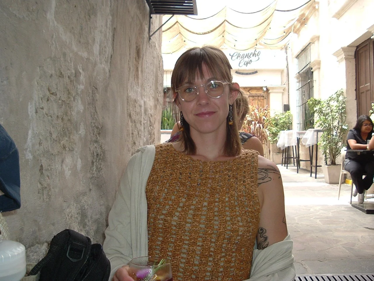 A woman with glasses and earrings sitting at an outdoor cafe, holding a drink. She has shoulder-length brown hair and is wearing a brown crochet top with a cream-colored jacket. The cafe has stone walls and table settings in the background.