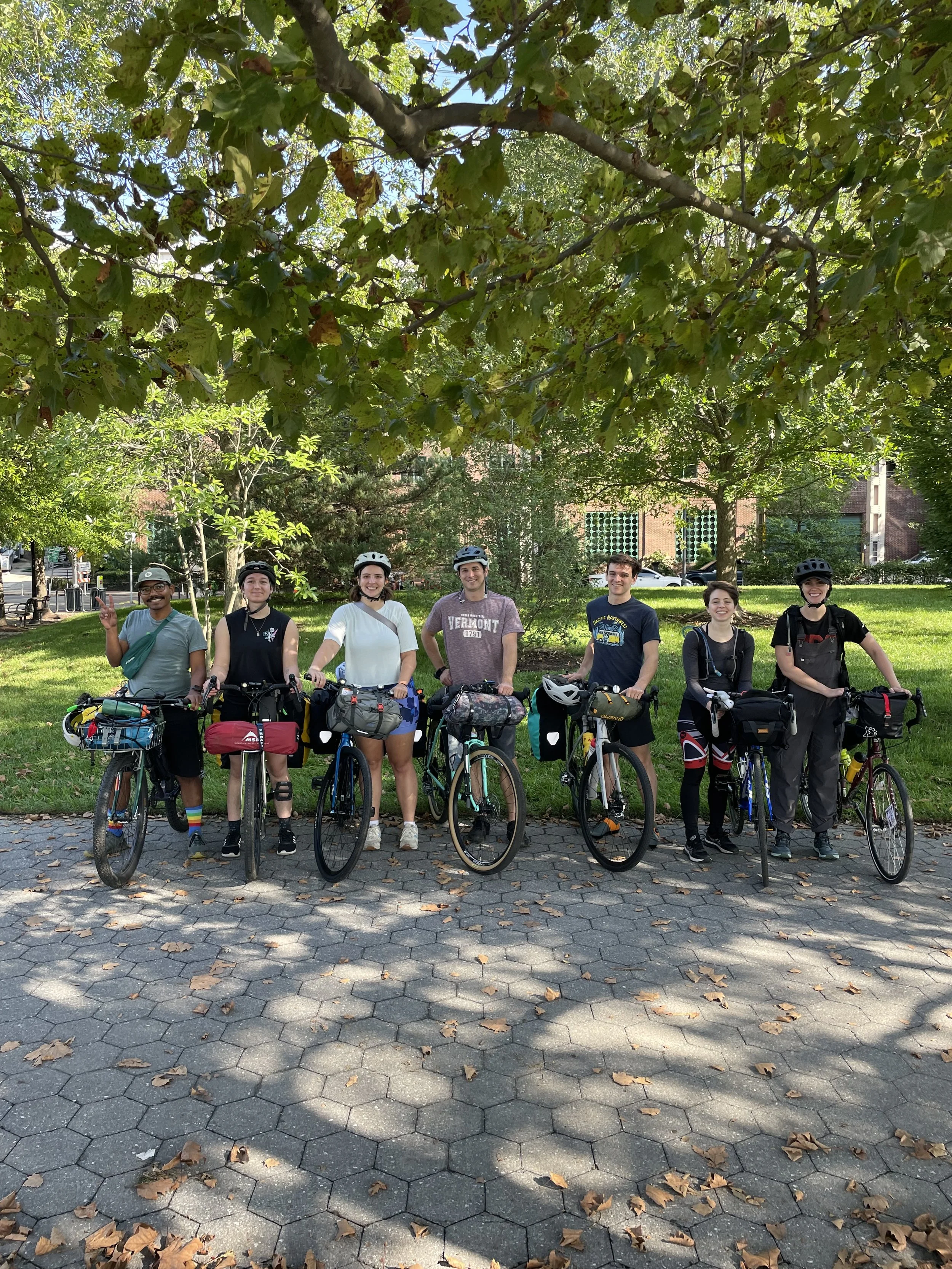 Group of seven people with bicycles standing outdoors on a paved area, smiling, under a large tree with green leaves.