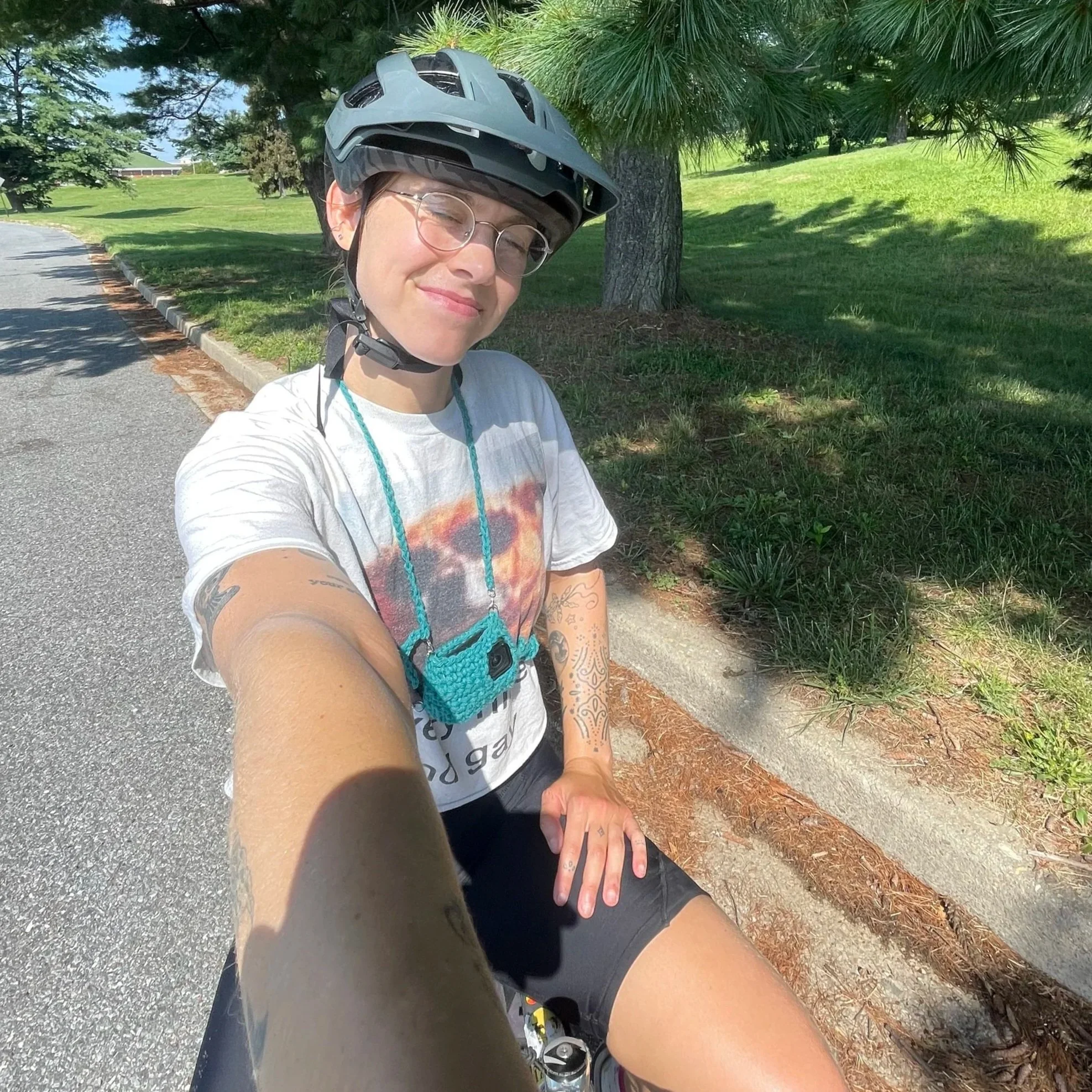 Young woman wearing a bicycle helmet, glasses, and a graphic t-shirt, taking a selfie during a bike ride on a sunny day.