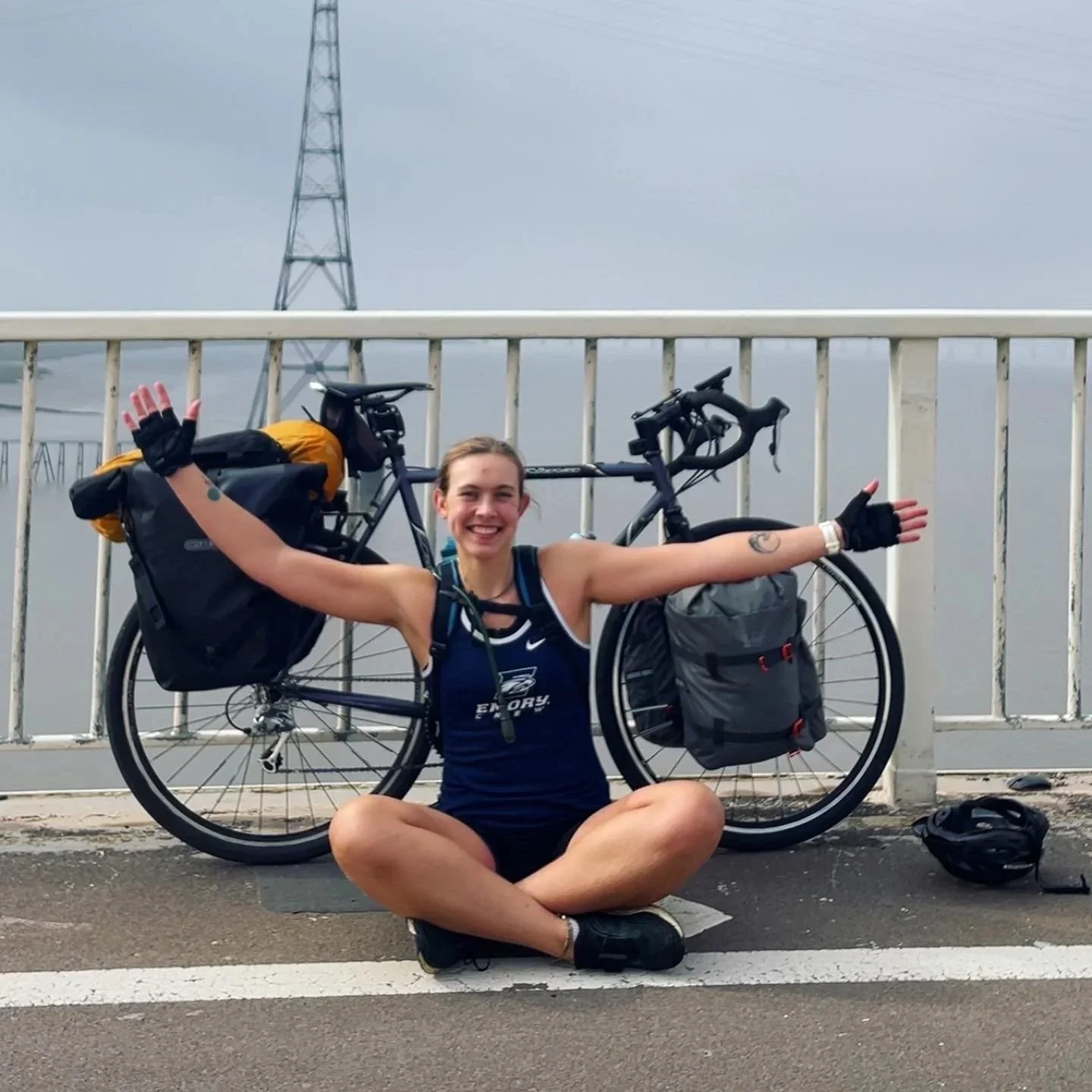 A woman sitting cross-legged on the road with a loaded bicycle behind her, arms open wide, smiling. The bicycle has multiple bags attached. There is a bridge with a railing and an electrical tower in the background.