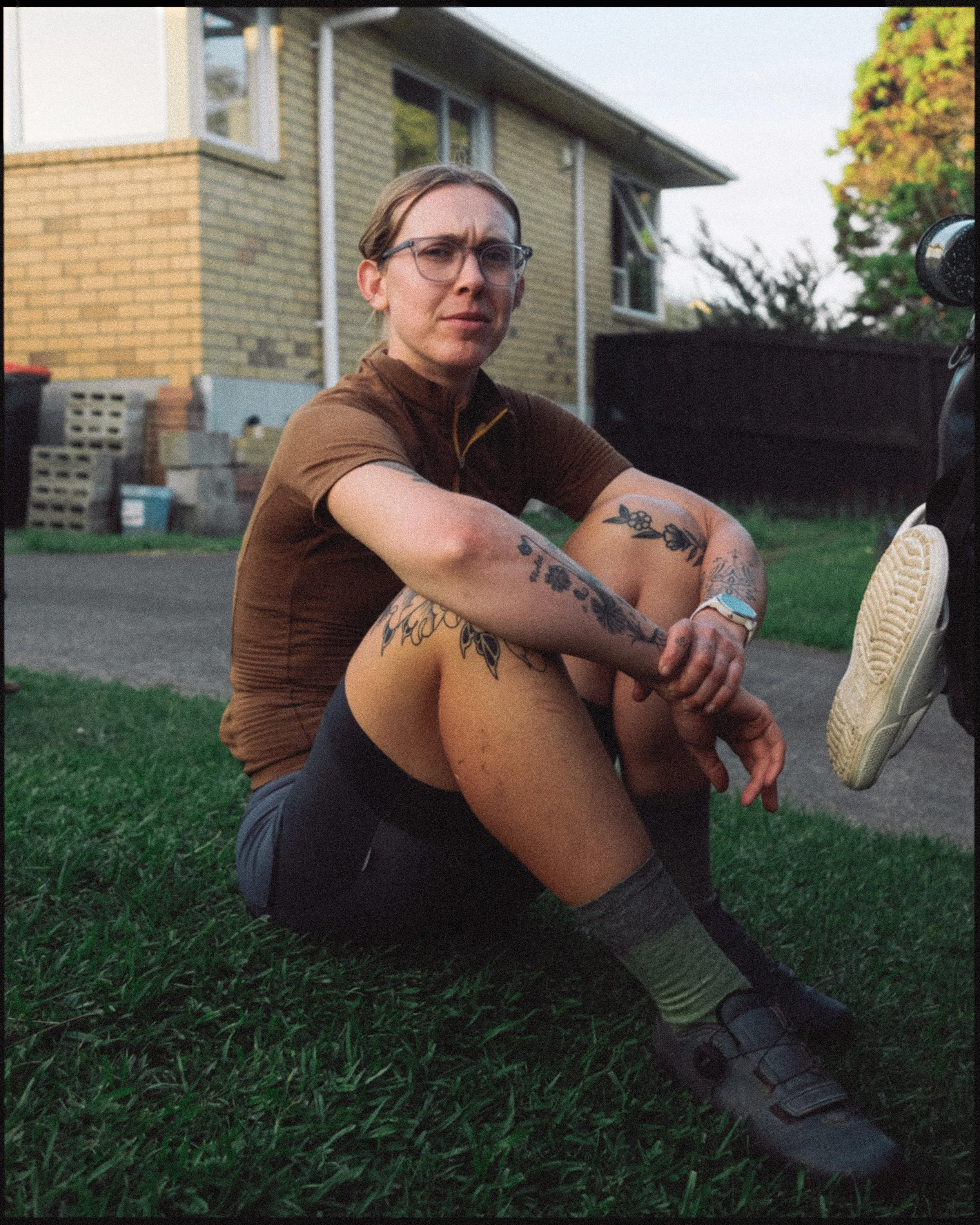 A woman with tattoos sitting on the grass outside a house, wearing glasses, a brown top, black shorts, and sandals.