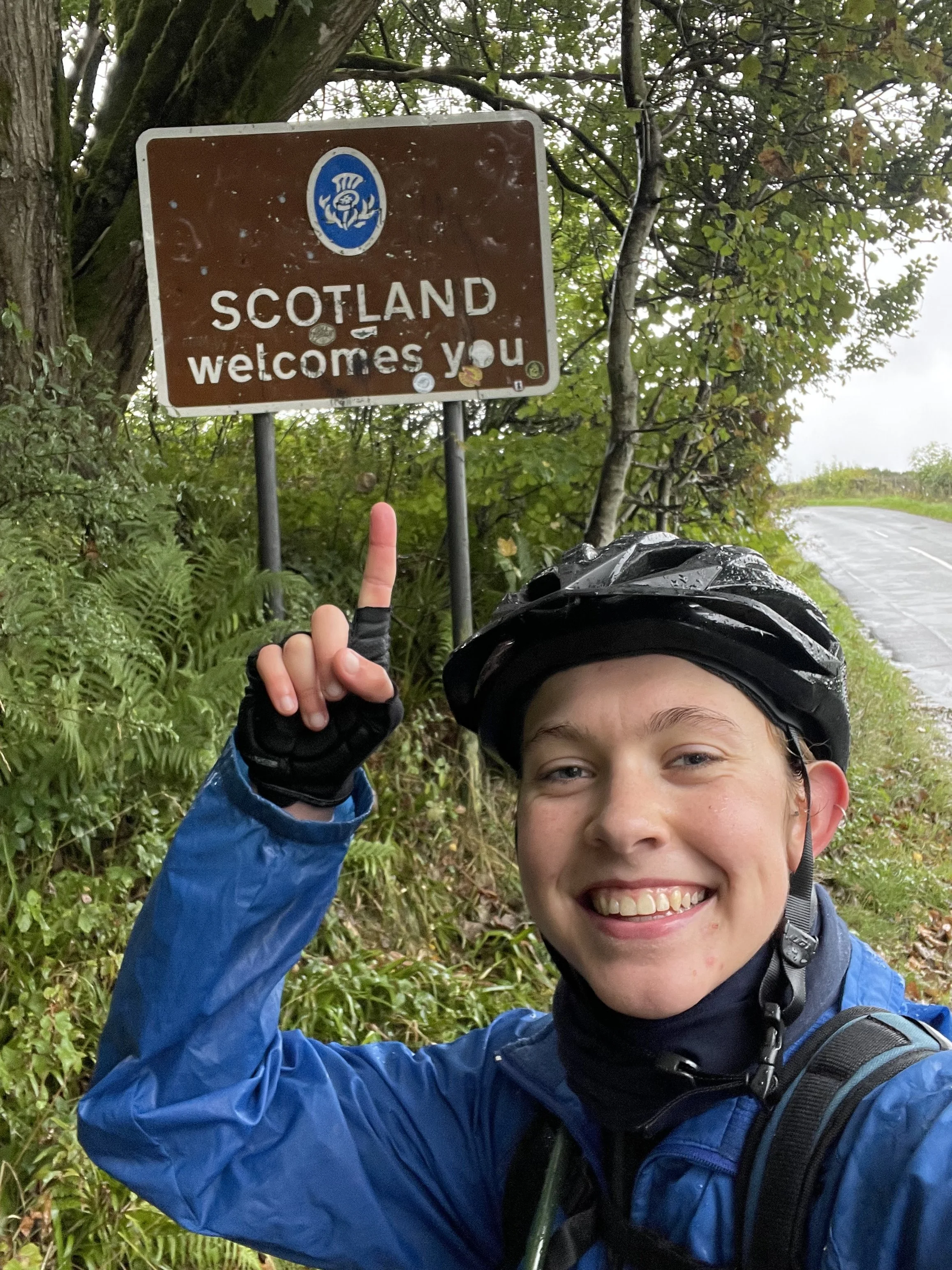 Smiling cyclist in blue jacket and black helmet pointing at a brown sign that says 'Scotland welcomes you' near a rural road surrounded by green foliage.