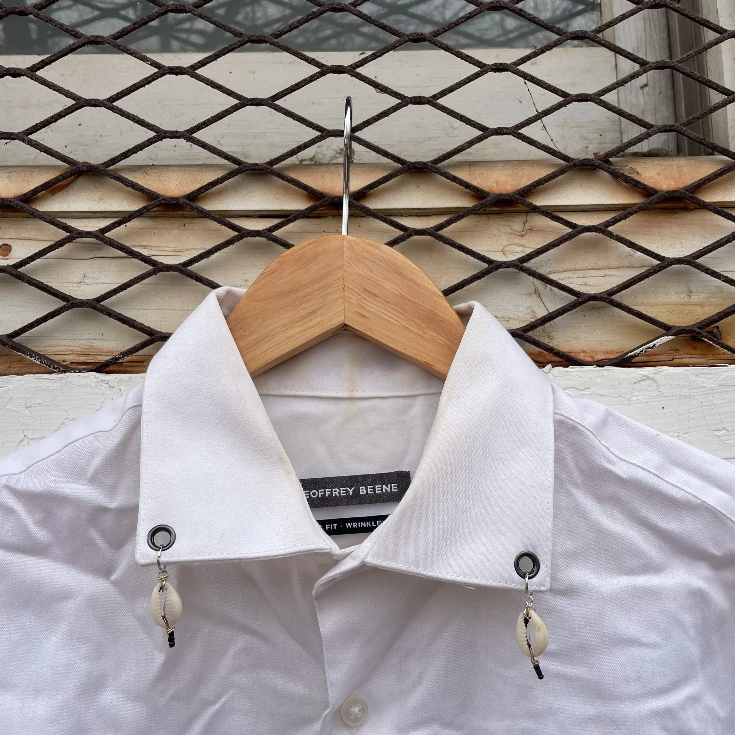 White button-up shirt hanging on a wooden hanger against a wall with a metal grid behind it.