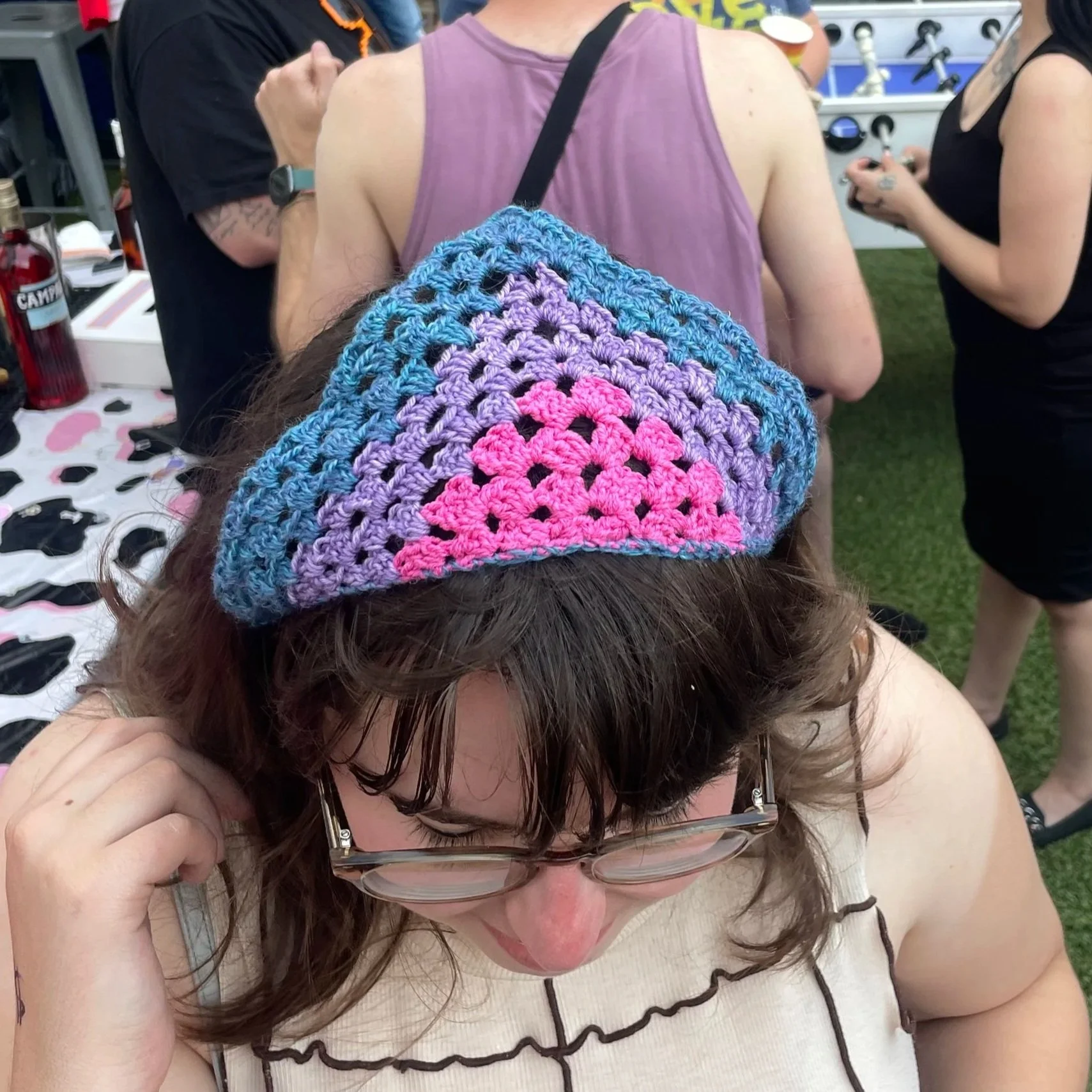 Person wearing a handmade, colorful, crocheted hat at an outdoor event, with others in the background