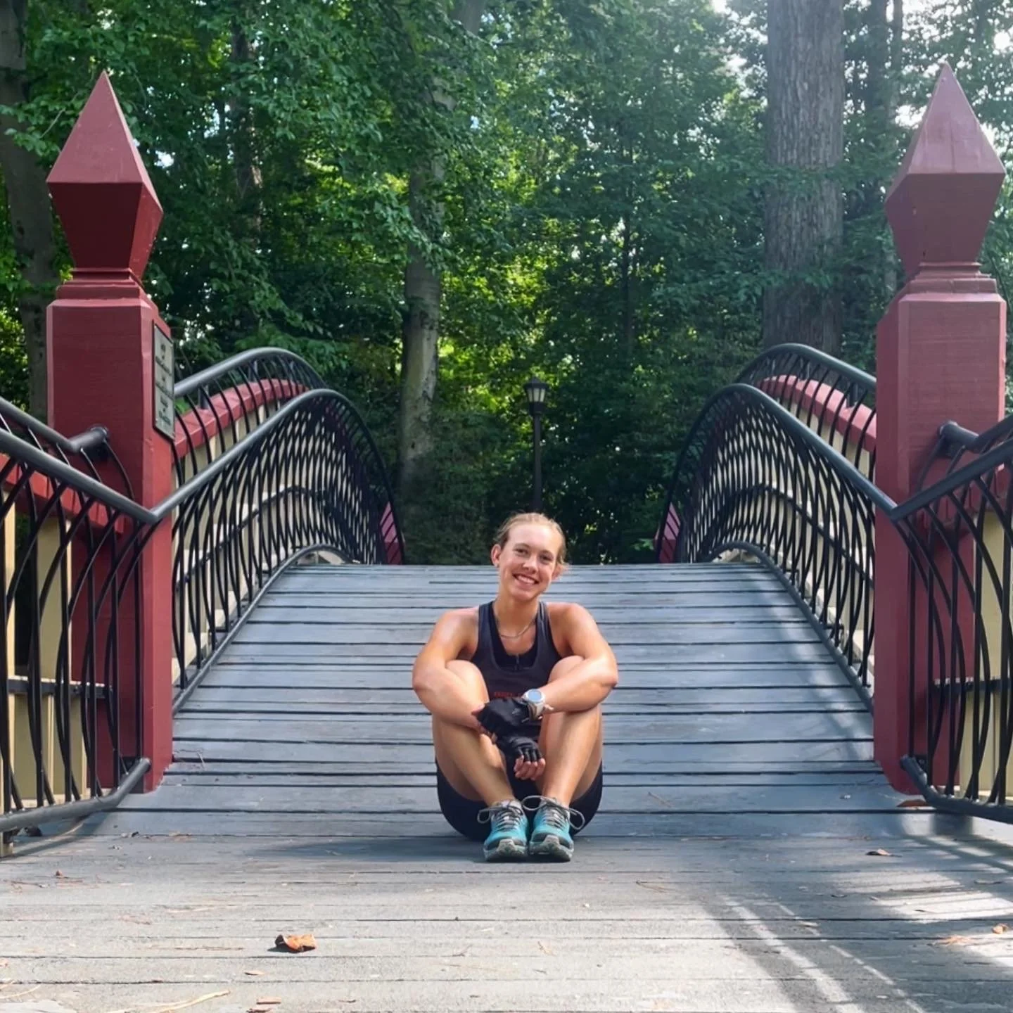 A woman sitting on a wooden bridge in a park, smiling at the camera, surrounded by green trees.