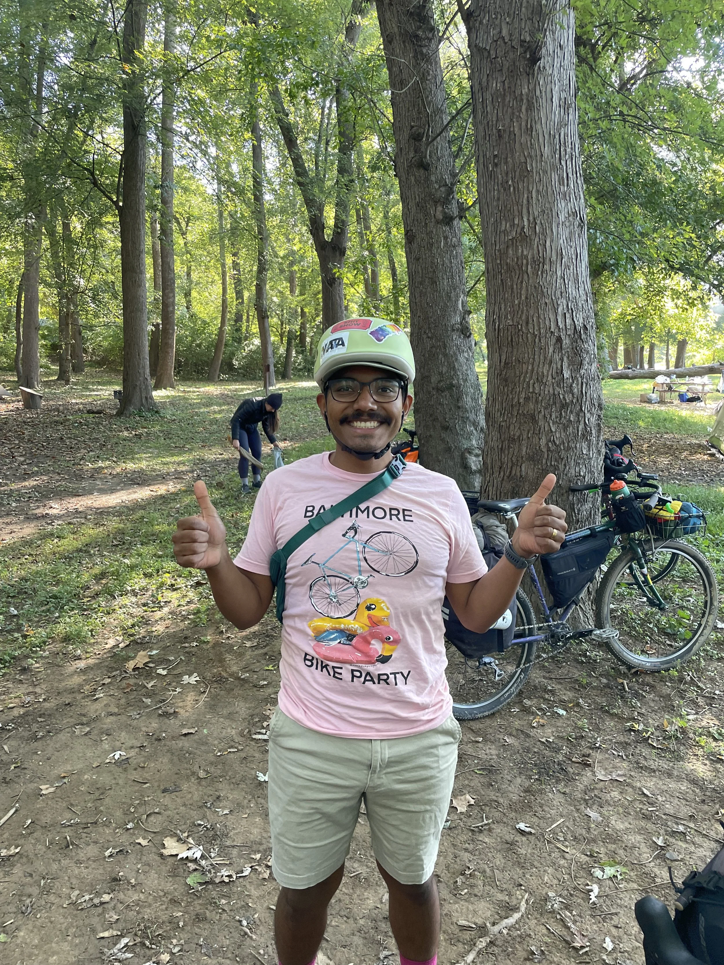 A smiling man wearing a pink bicycle-themed t-shirt, shorts, glasses, and a helmet, standing in a wooded park giving two thumbs up. There are bicycles and another person in the background.