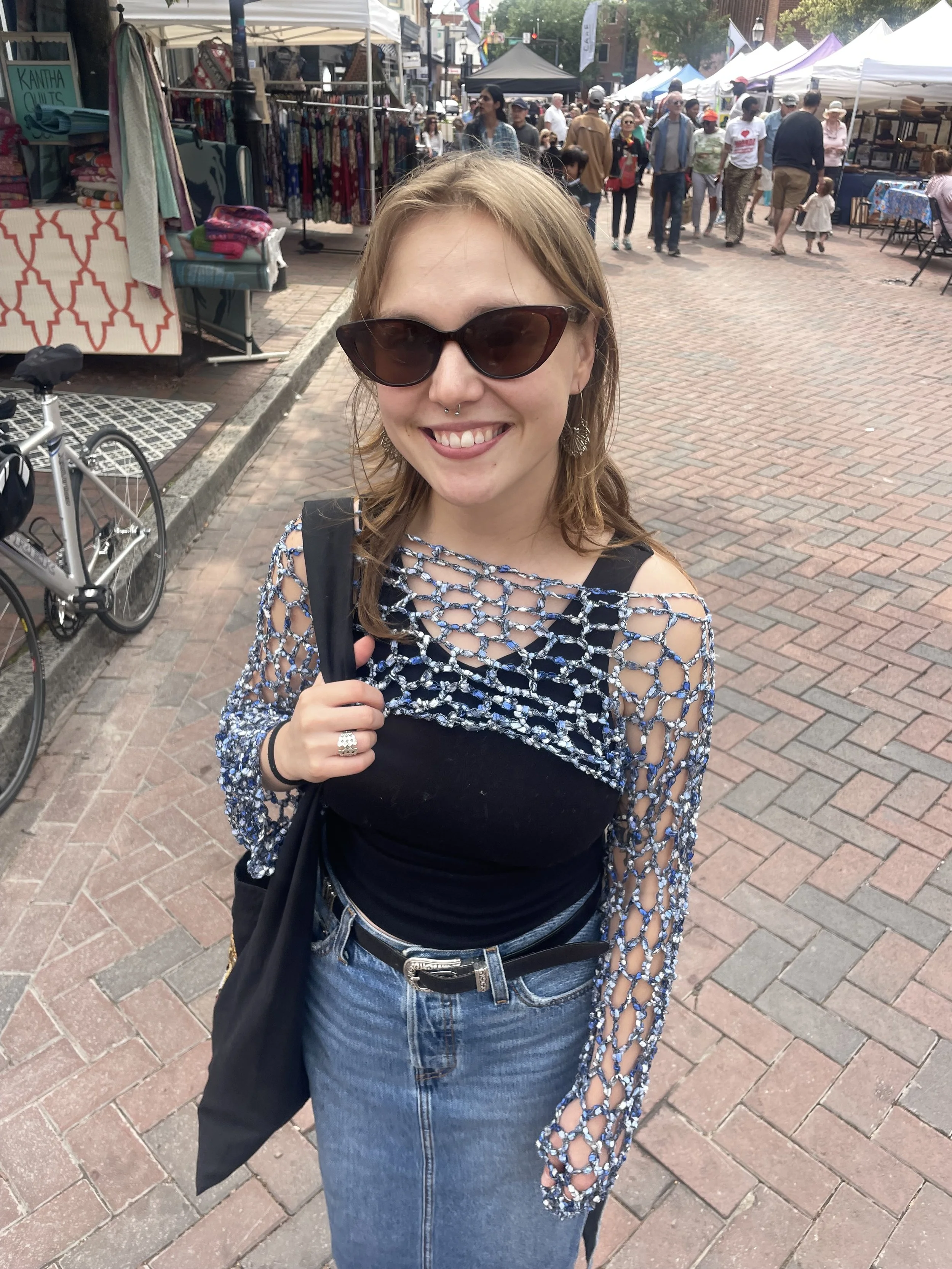 A young woman with light brown hair wearing sunglasses, a black top with a plastic netted overlay, and blue jeans standing at an outdoor market with many people, tents, and vendor stalls in the background.