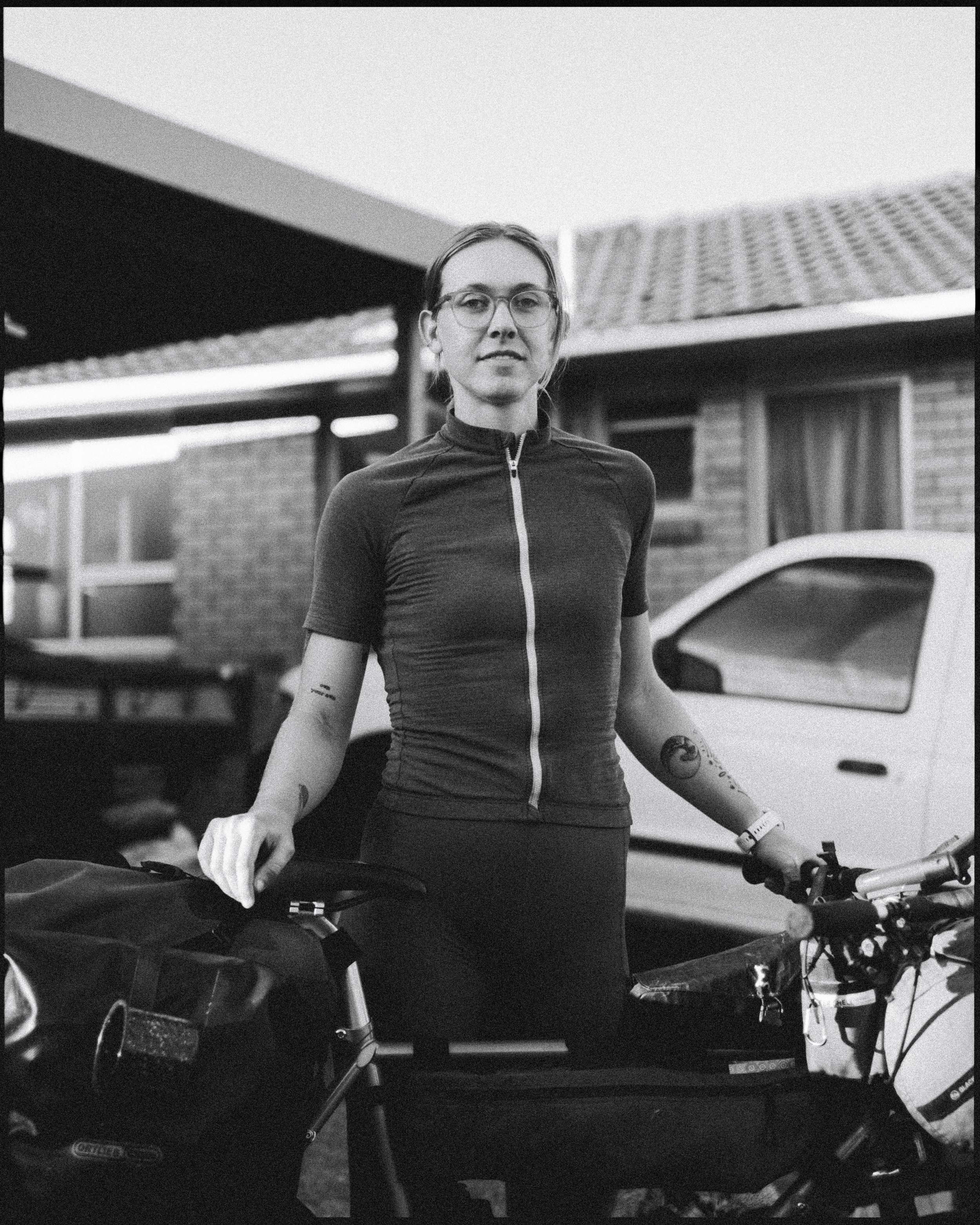 A woman with glasses standing outdoors with a bicycle, in front of a brick house and car, wearing a athletic shirt.