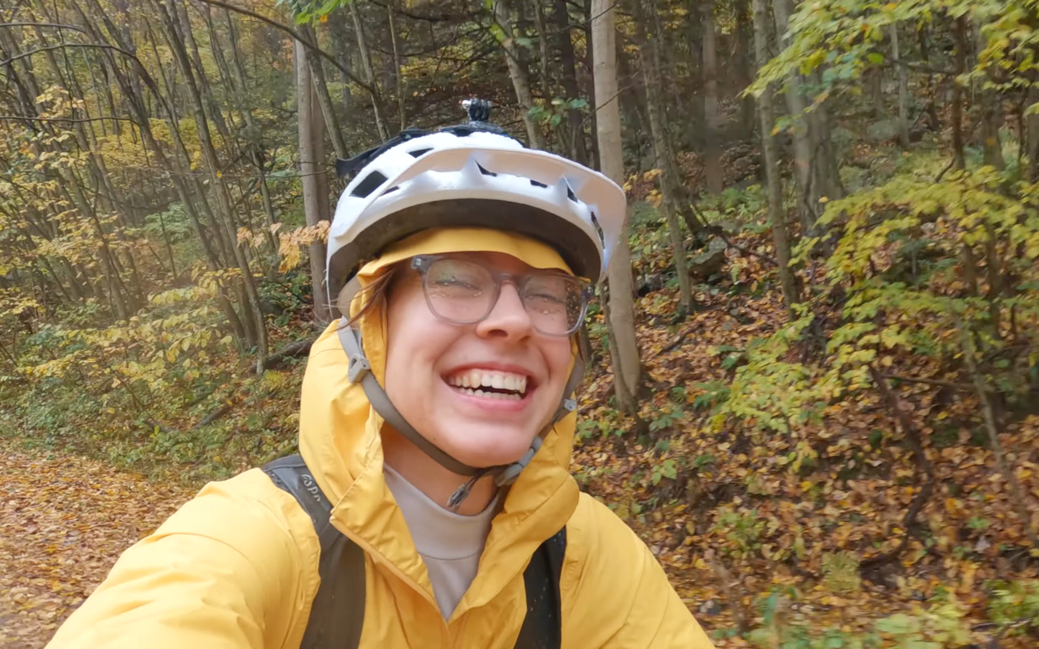 A person in a yellow jacket and white helmet smiling while mountain biking in a forest with fall-colored leaves.