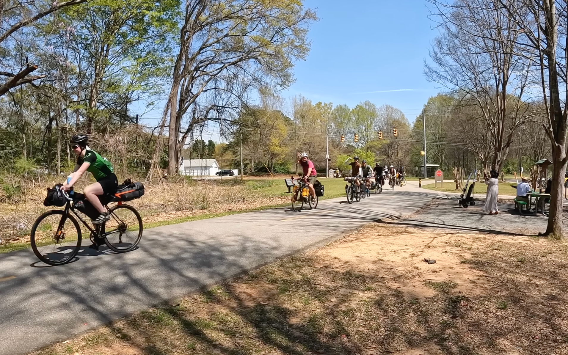 Multiple cyclists riding on a paved trail in a park on a sunny day, with trees and benches along the path, and pedestrians sitting and standing nearby.