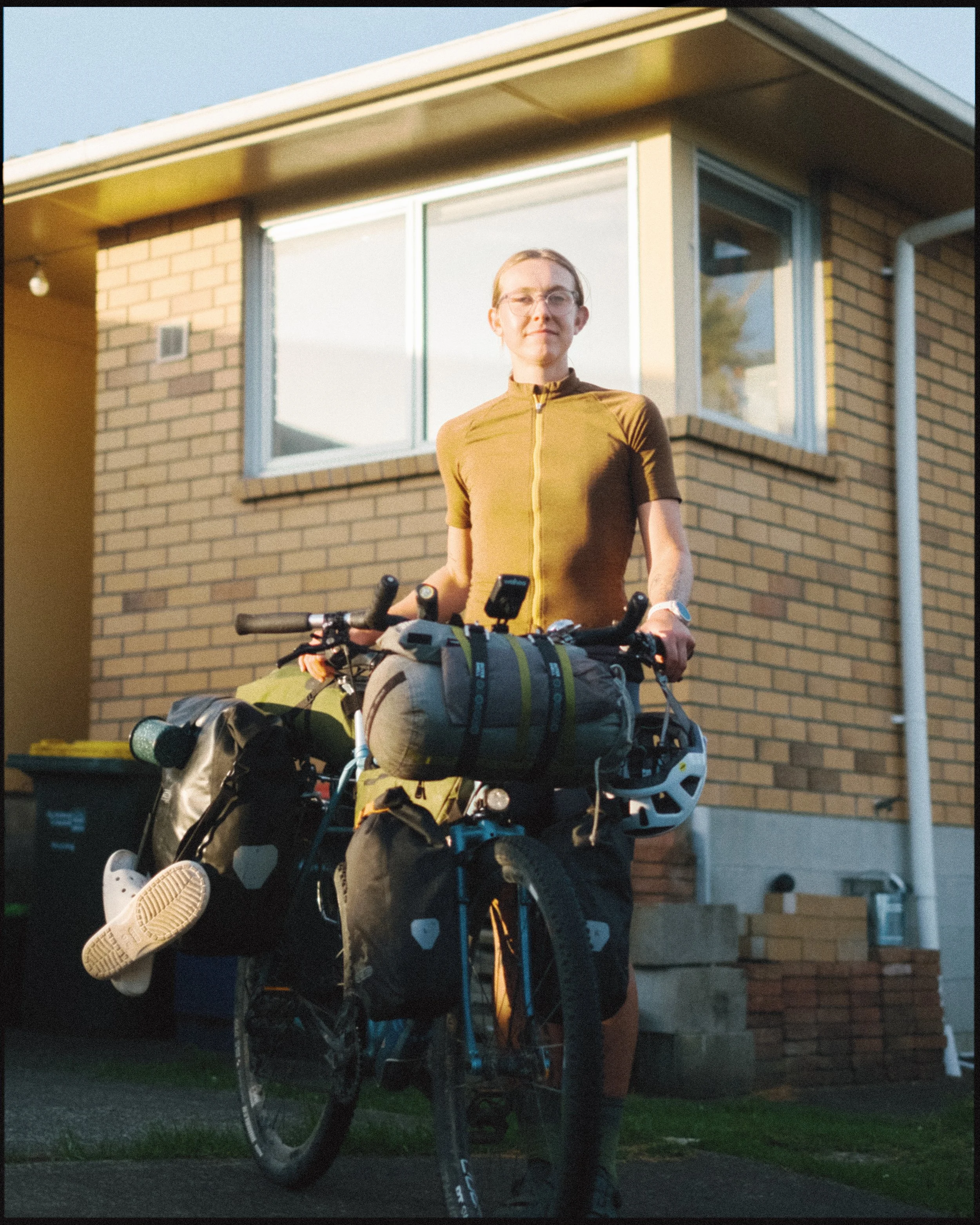 Person standing outside a brick house with a loaded touring bicycle, wearing a yellow-brown cycling jersey, glasses, and a watch.