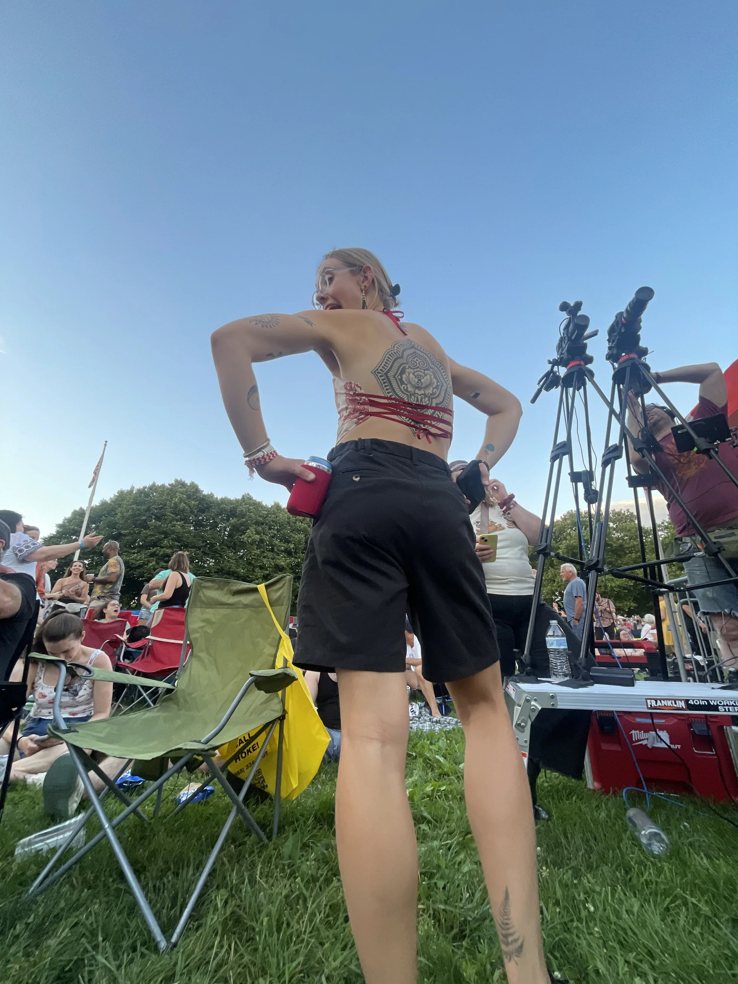 A woman standing outdoors at a crowded event, wearing a red and beige top and black shorts, with tattoos visible on her arms, back, and legs. She is holding a red cup and appears to be in conversation, with cameras and other people around.