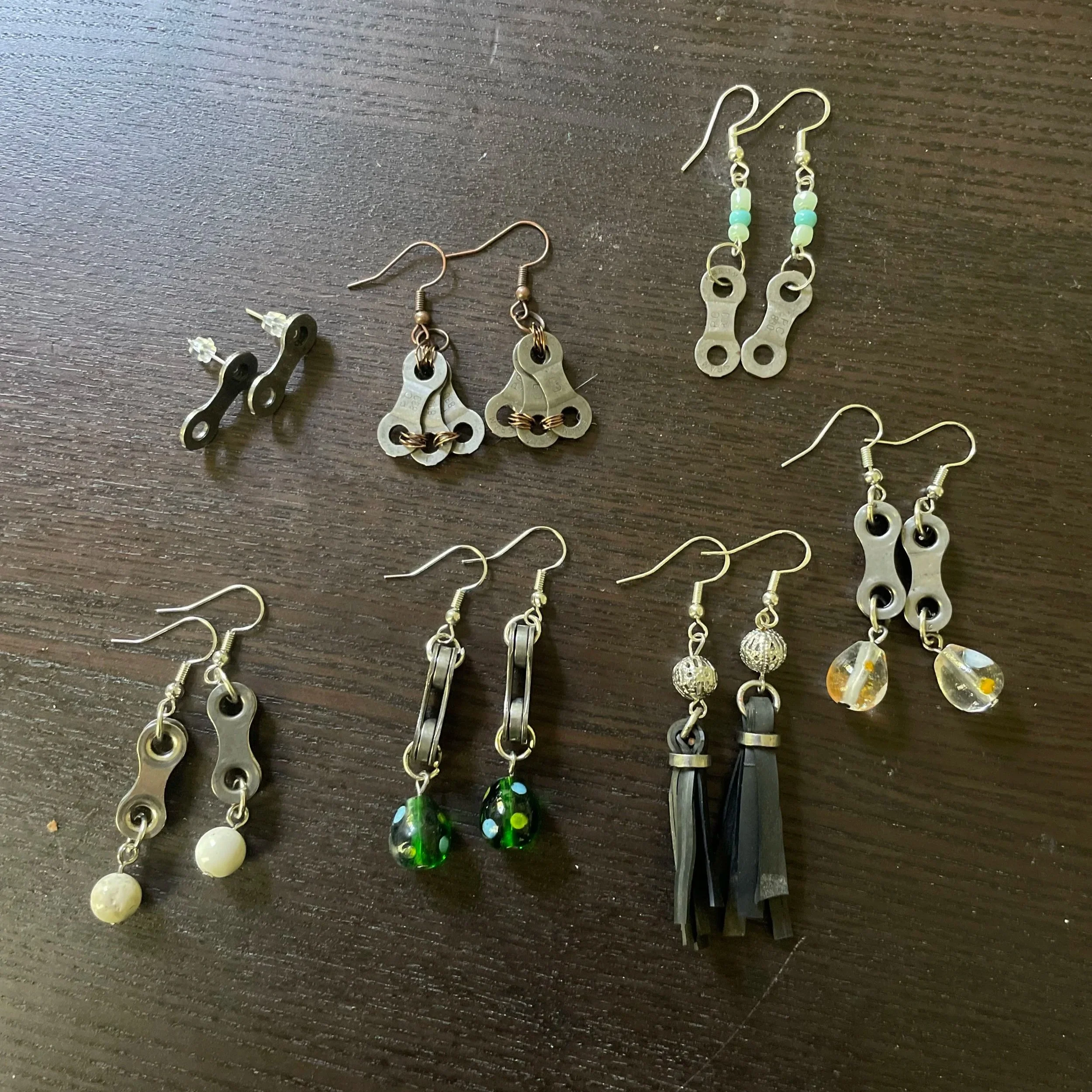 Six pairs of earrings, including bike chain style, beaded, and dangle designs, arranged on a dark wooden surface.
