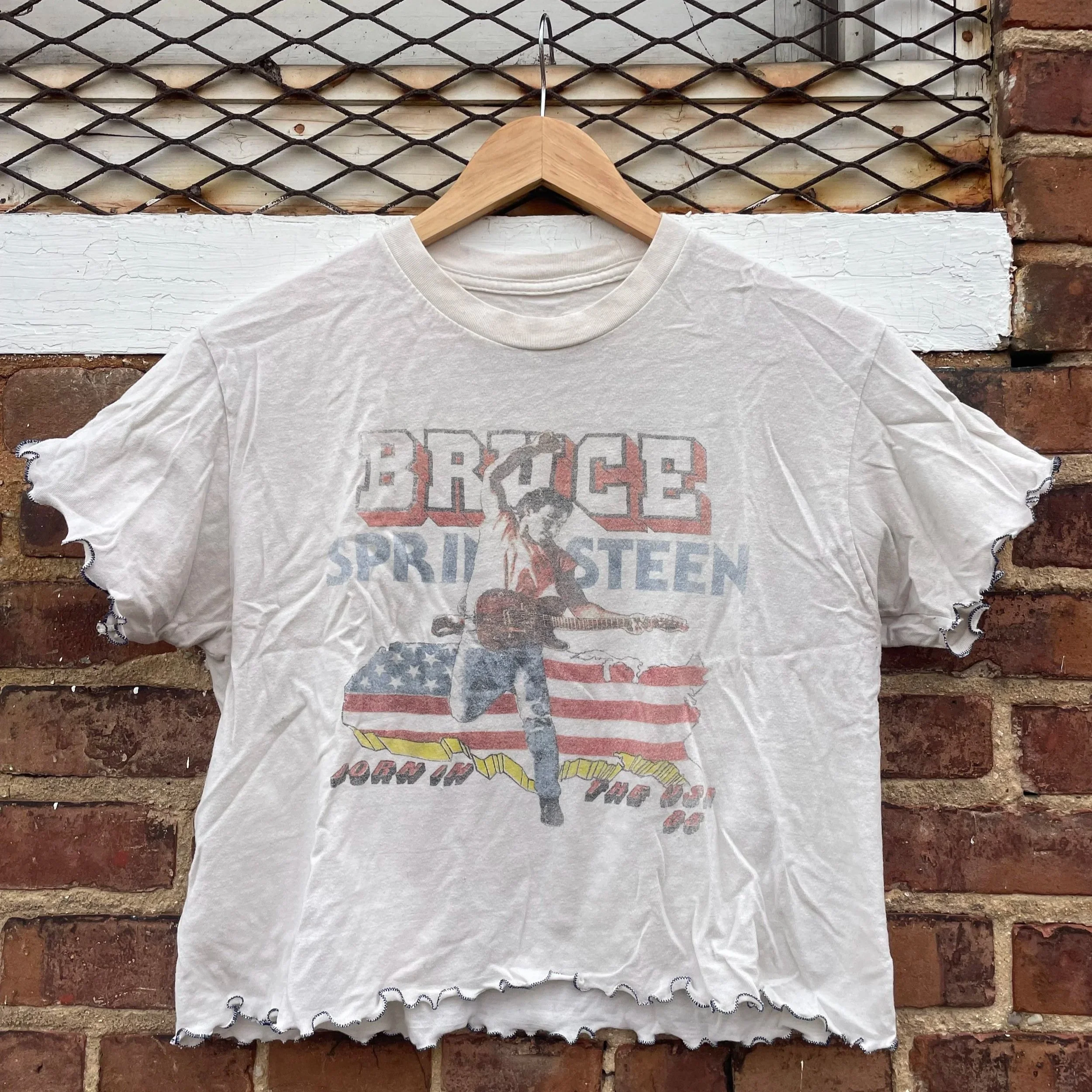 A white t-shirt with a graphic of Bruce Springsteen playing guitar in front of an American flag background, with text that reads "BRUCE SPRINGSTEEN" and some additional smaller text.