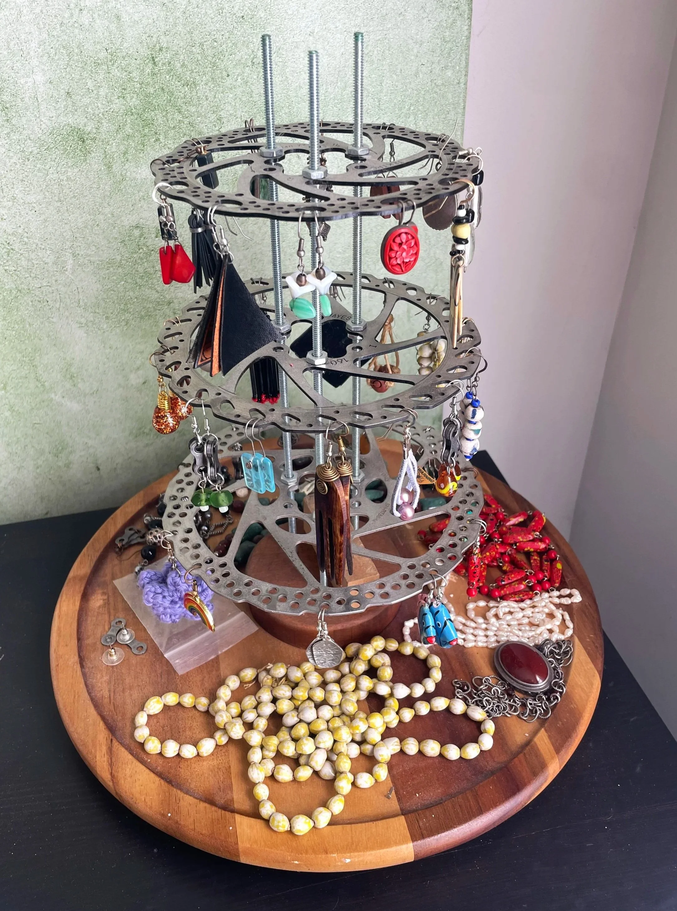 A jewelry display stand with multiple tiers holding various earrings and necklaces, placed on a wooden round table with extra jewelry pieces surrounding it.