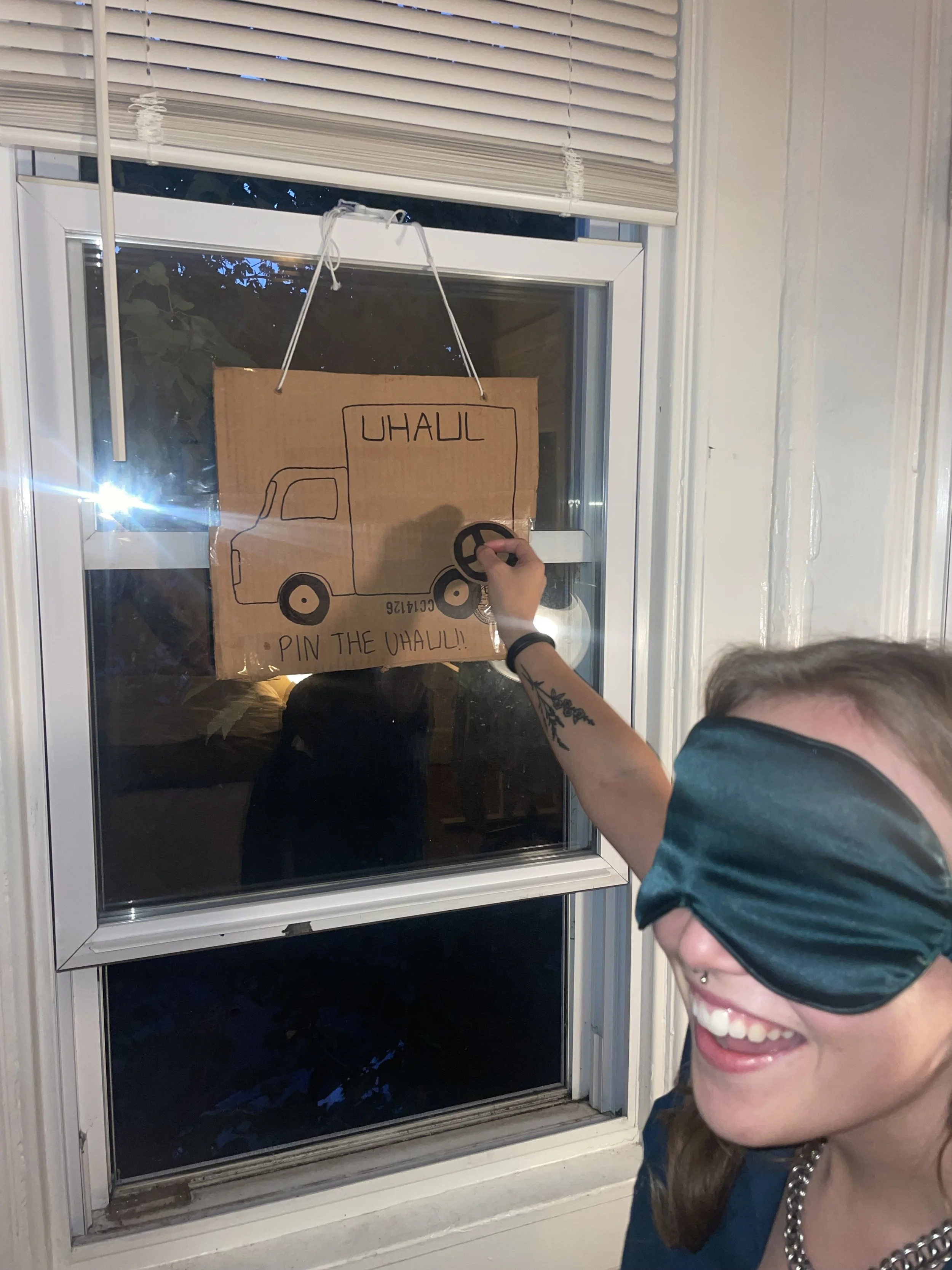 A woman with a mask on, smiling, playing a pin-the-wheel game on a cardboard U-Haul sign on a window.