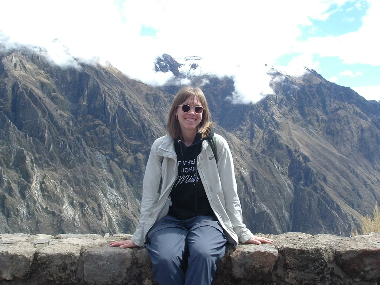 A woman with sunglasses sitting on a stone wall outdoors in front of towering mountain peaks with some clouds and a partly cloudy sky.