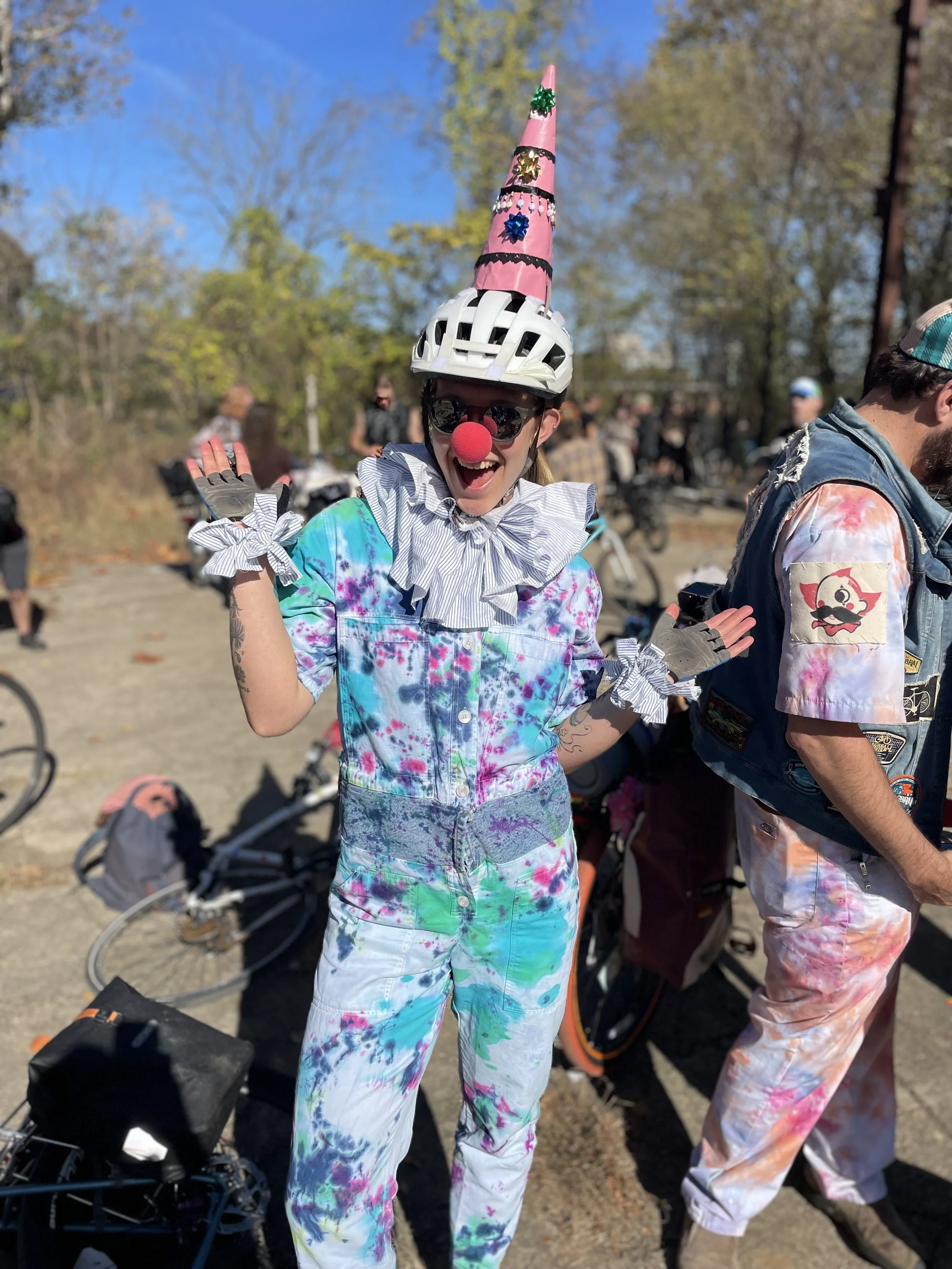 Person dressed as a clown with a rainbow tie-dye outfit, red clown nose, and a birthday hat, smiling and waving outdoors with bicycles and other people in the background.