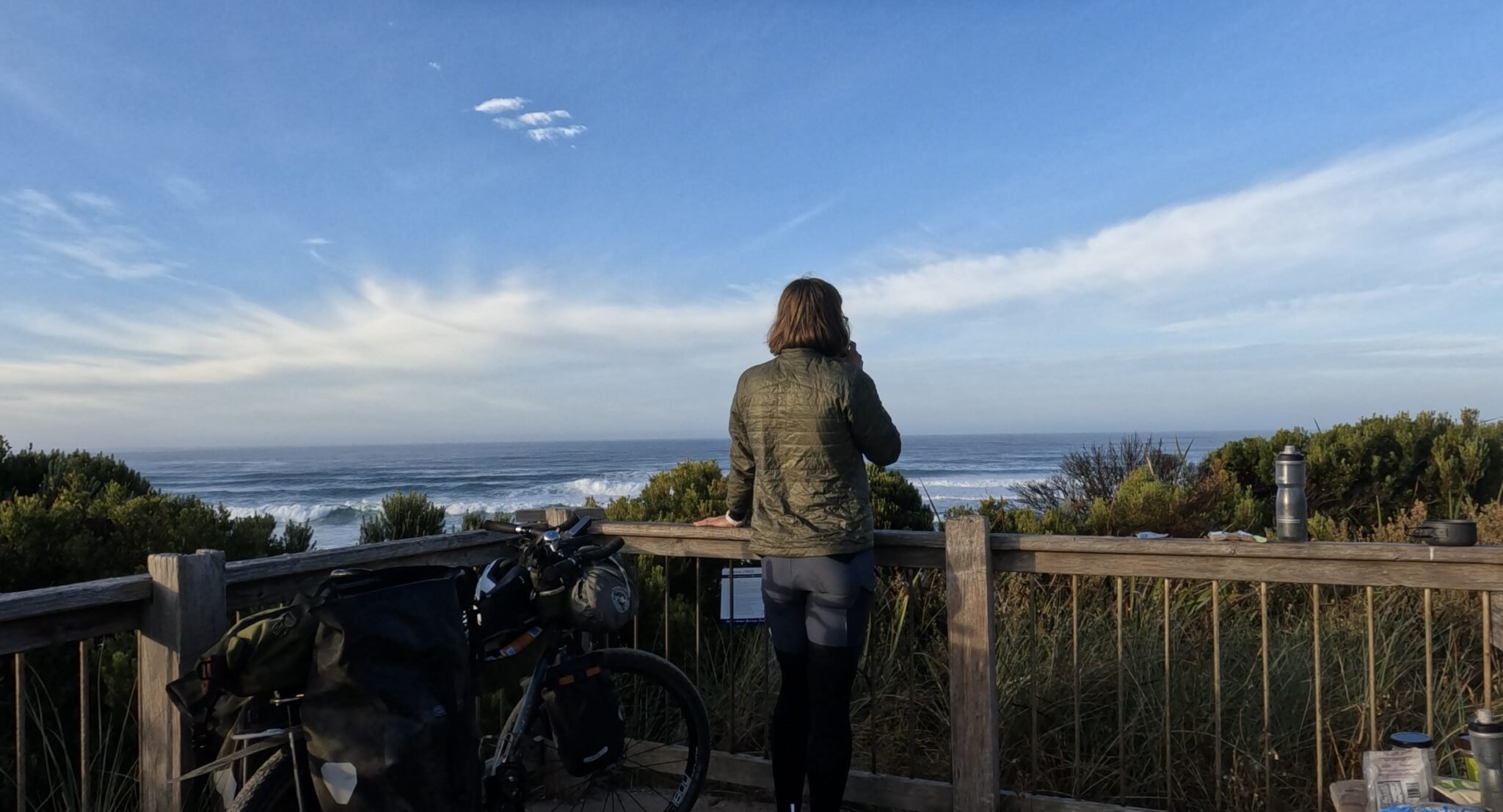 A person with short hair, wearing a green jacket and black pants, standing on a wooden deck overlooking the ocean, with a bicycle and water bottles nearby