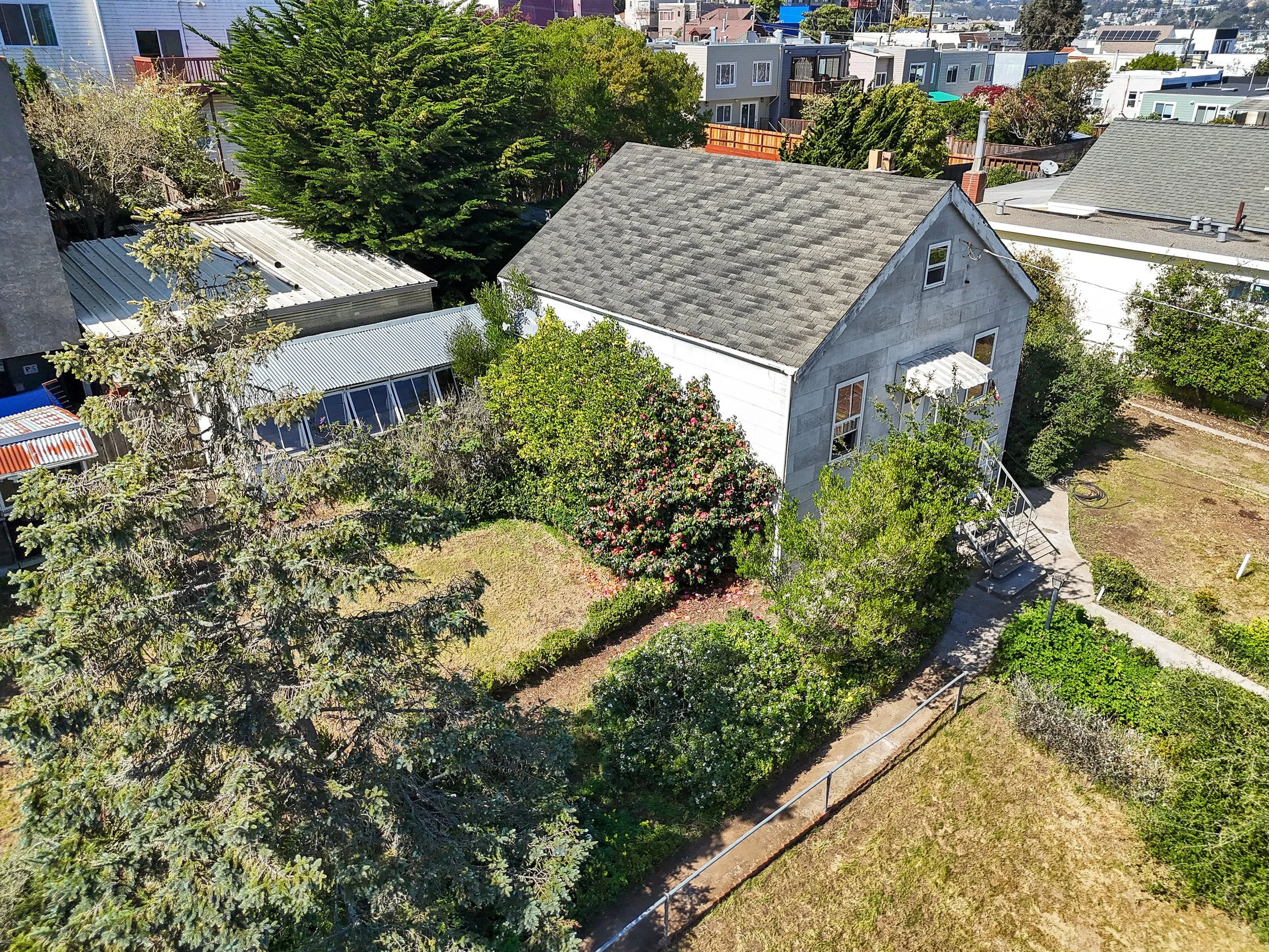 An aerial view of a backyard with trees, bushes, and a two-story house with a gray roof and light exterior walls. The yard has a pathway, a small lawn, and surrounding neighboring houses.