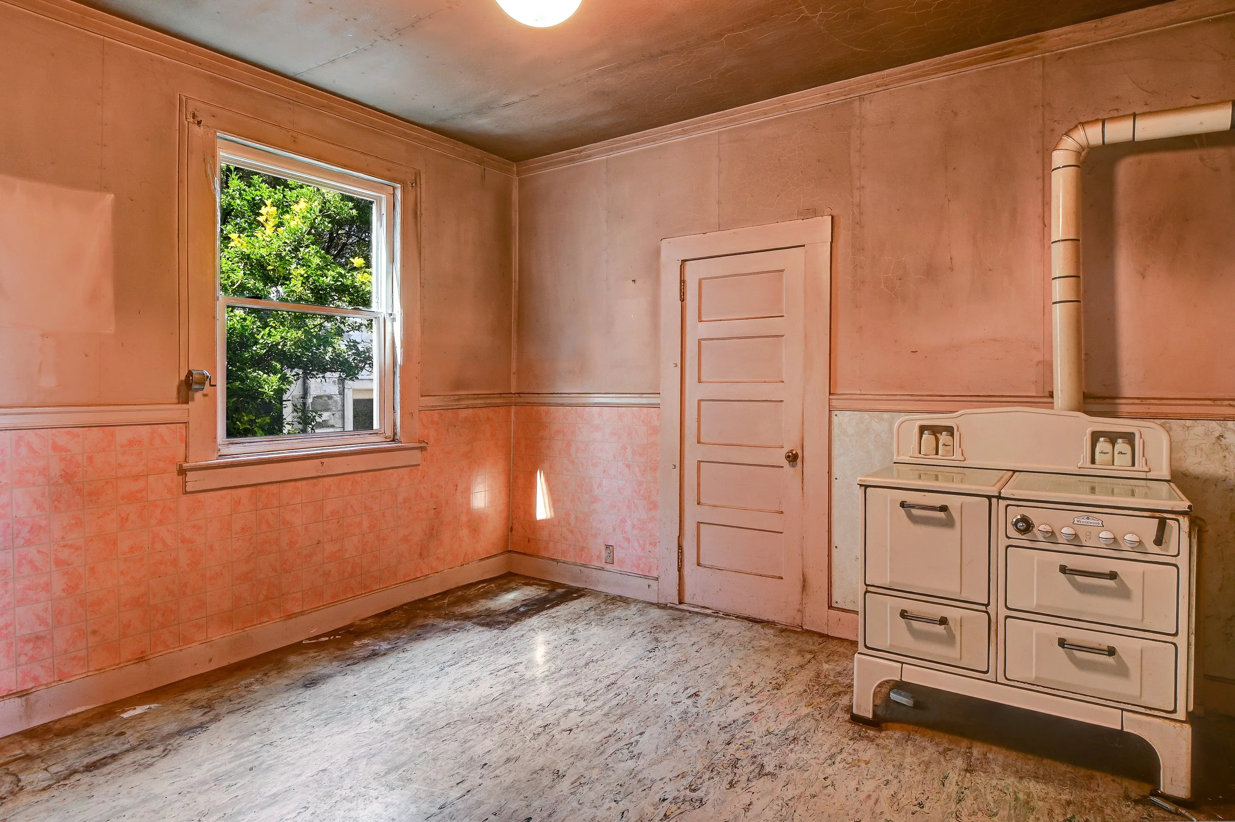 Old, empty kitchen with pink walls and a window showing green foliage outside. A vintage white stove is in the corner, and the floor is worn and stained.